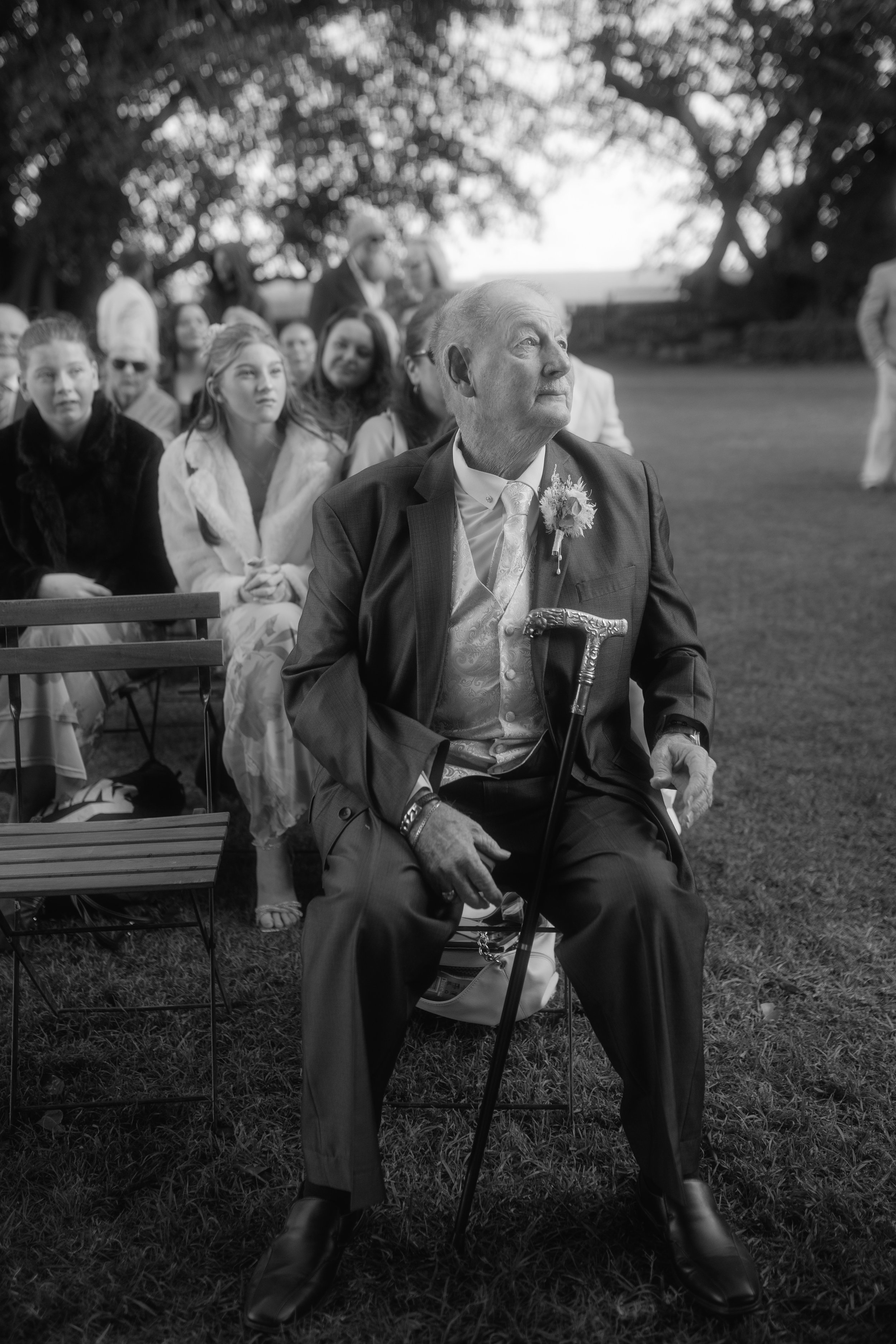 An elderly man in a suit and bow tie sitting on a chair outdoors, holding a cane, during a formal event. In the background, several guests, including women and men, are sitting and standing under large trees, at what appears to be a garden wedding ce