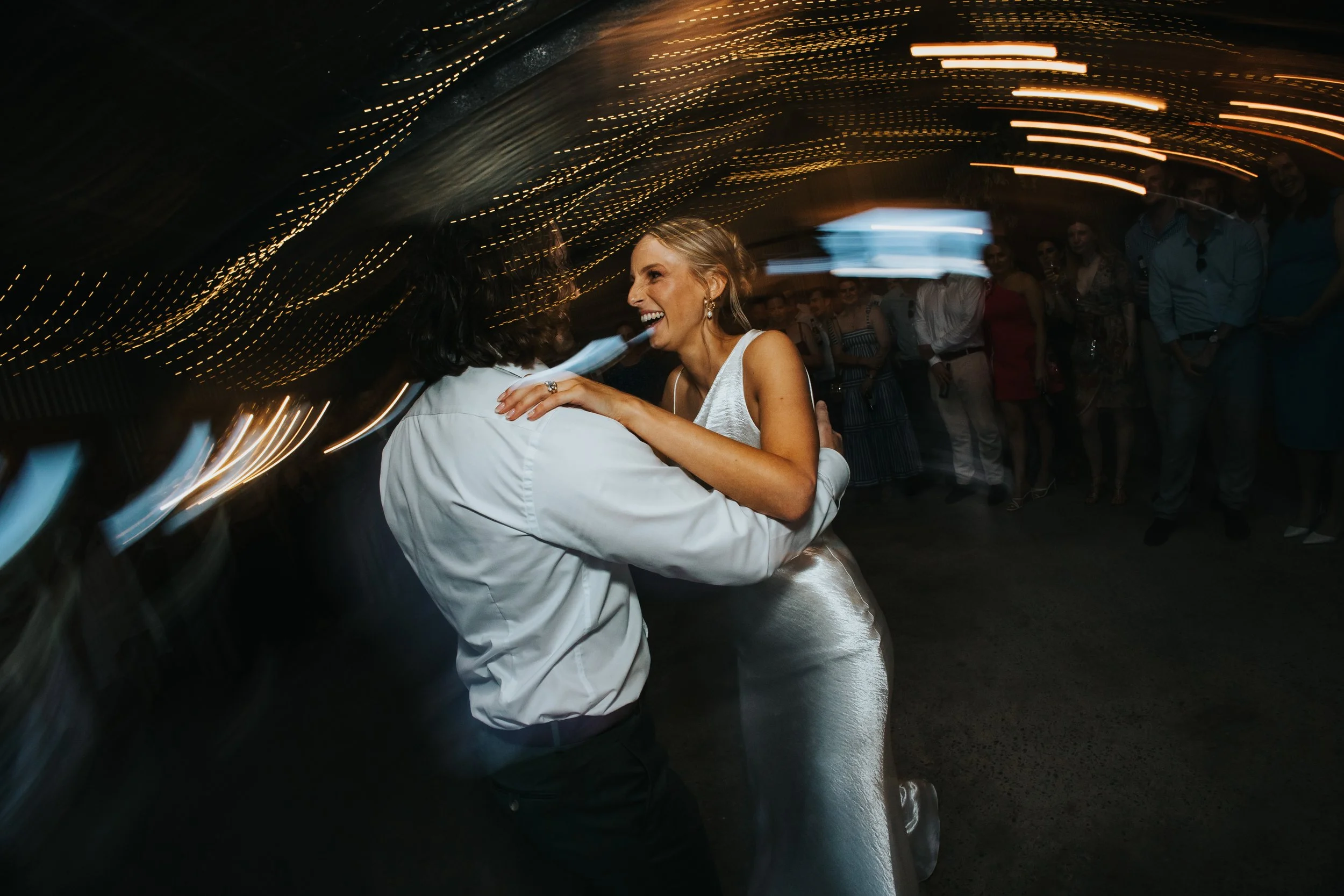 A woman smiling and dancing with a partner on a dance floor at night, surrounded by other guests, with string lights overhead creating a festive atmosphere.