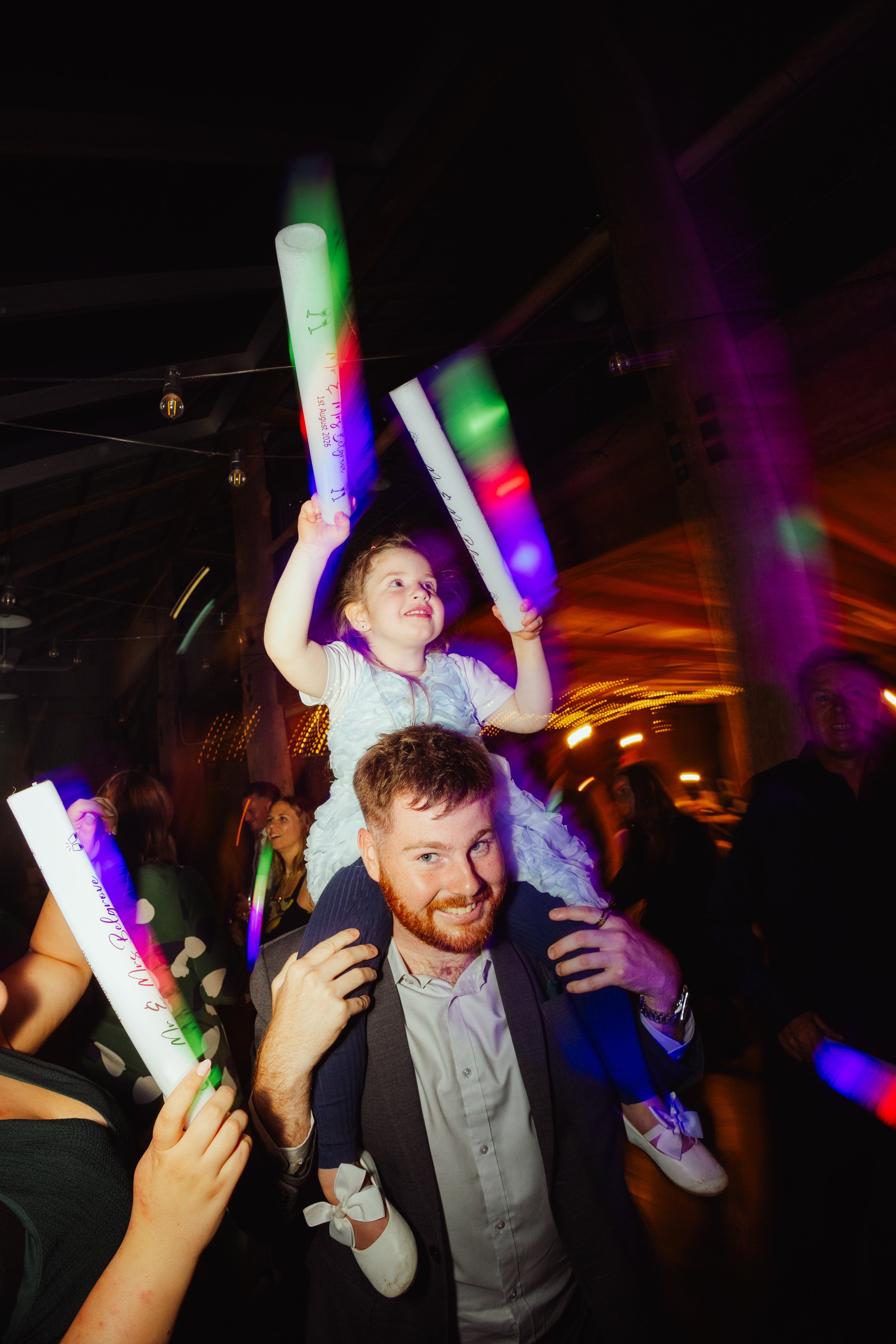 A joyful man with a beard and a woman with a child on his shoulders celebrating at a party or event with colorful lights and glow sticks.
