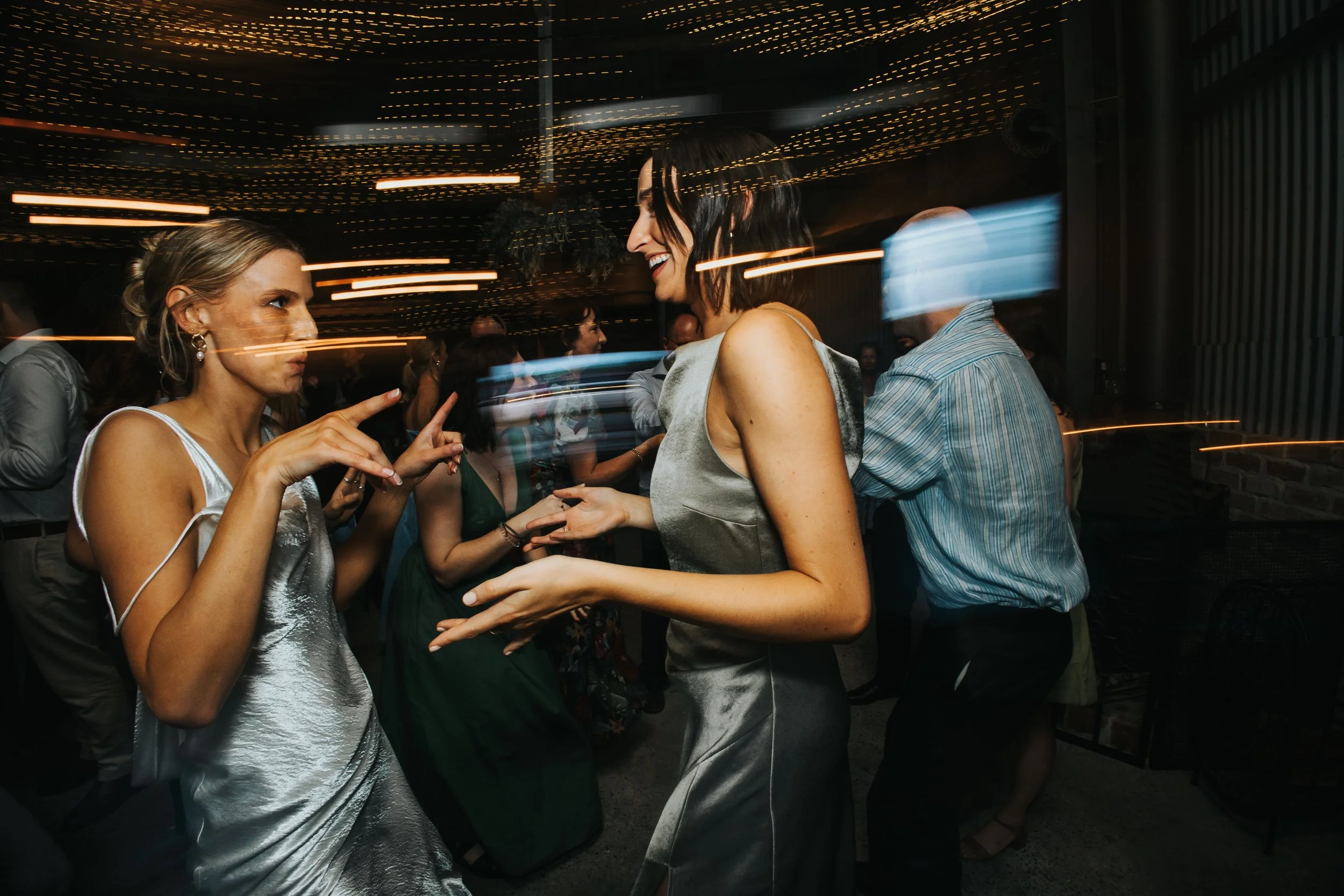 Two women dance and smile at a party or event with string lights overhead. Other people are visible in the background.