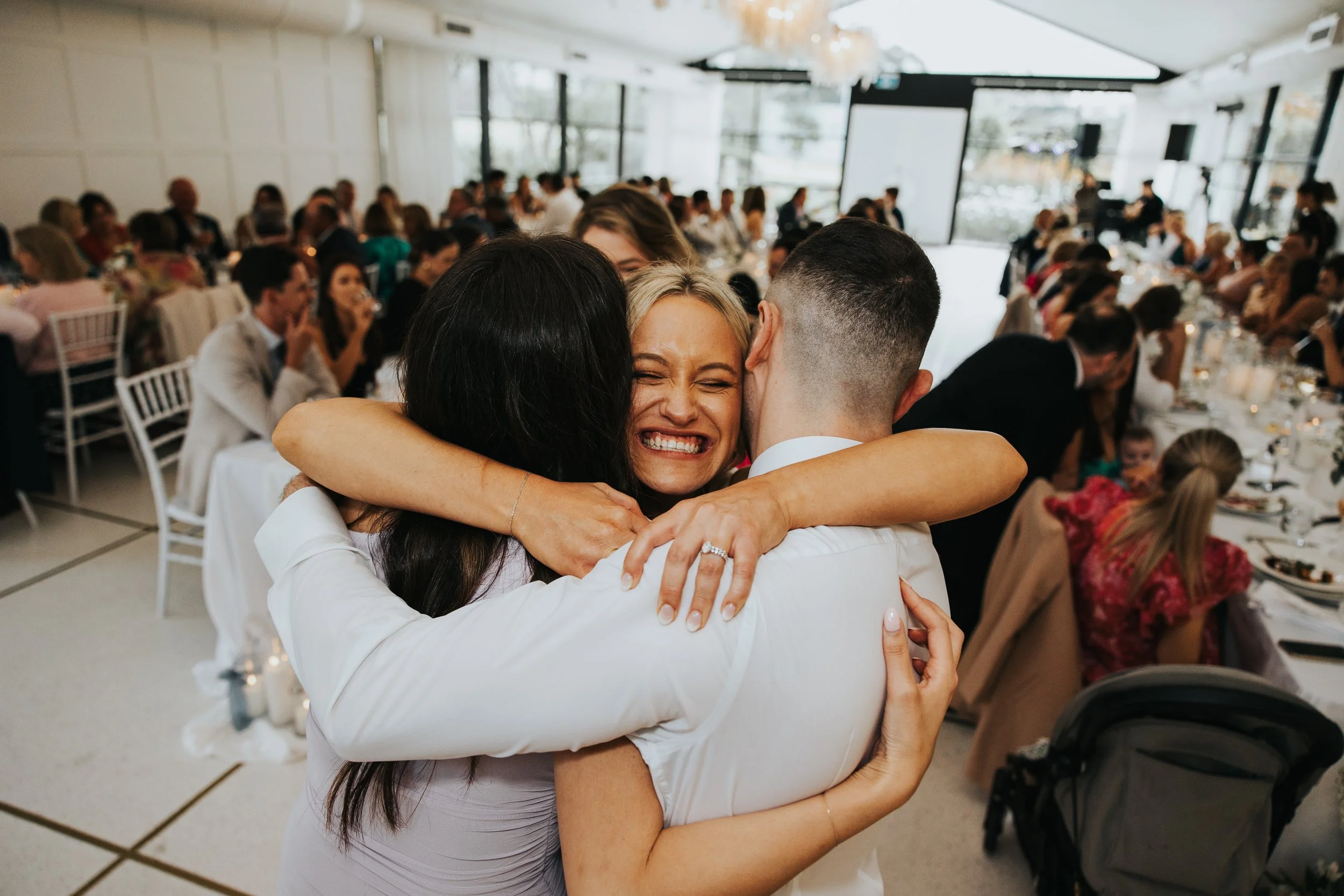 People embracing at a celebration or event in a well-lit banquet hall.