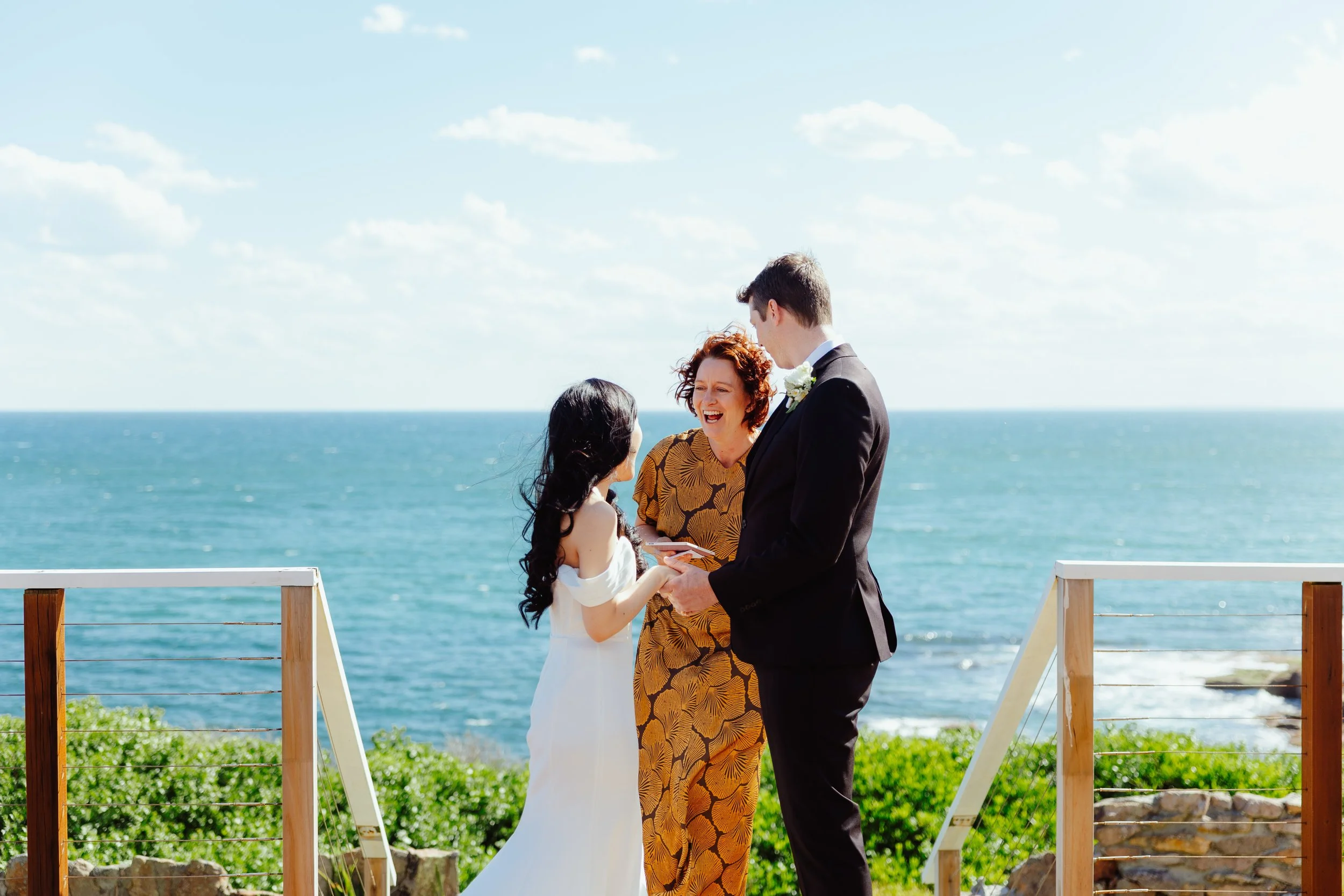 A couple getting married outside near the ocean, with an officiant officiating, under a partly cloudy sky.