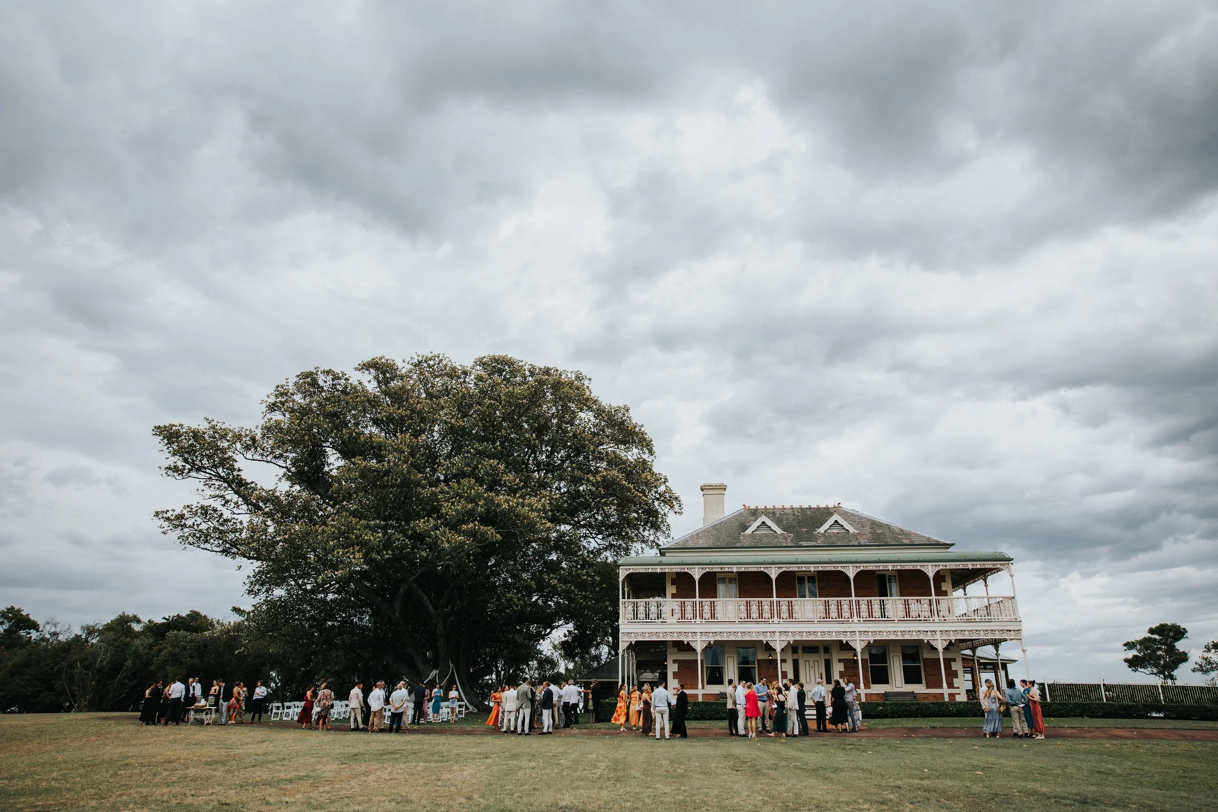 A large tree with green leaves stands in front of a Victorian-style house with a large porch, many windows, and white trim. There are numerous people gathered on the lawn, some standing and some sitting, dressed in semi-formal attire. The sky is over