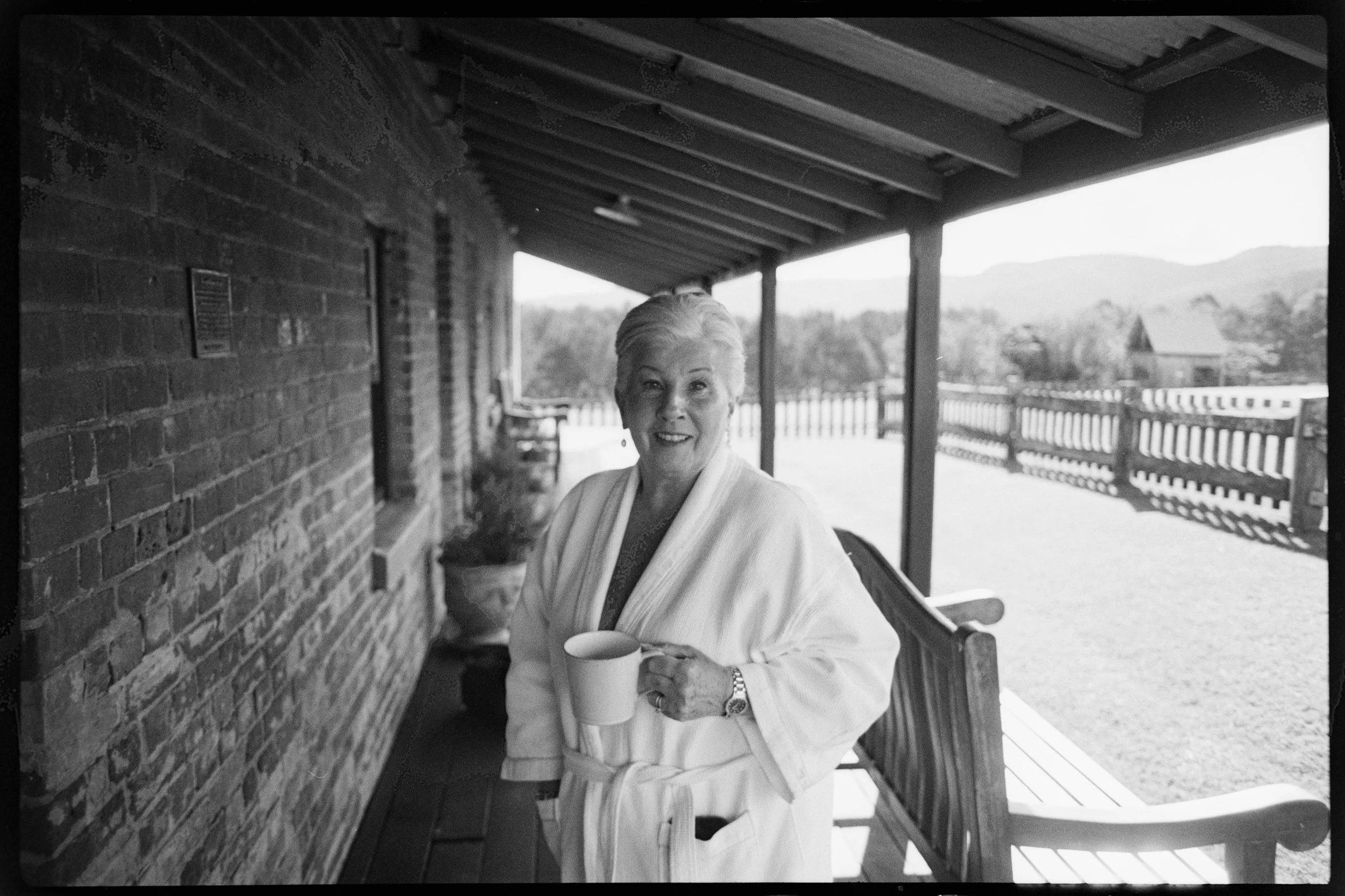 An elderly woman with short, white hair smiling and holding a coffee mug. She is standing on a porch with a wooden railing, next to a brick wall, with countryside in the background.