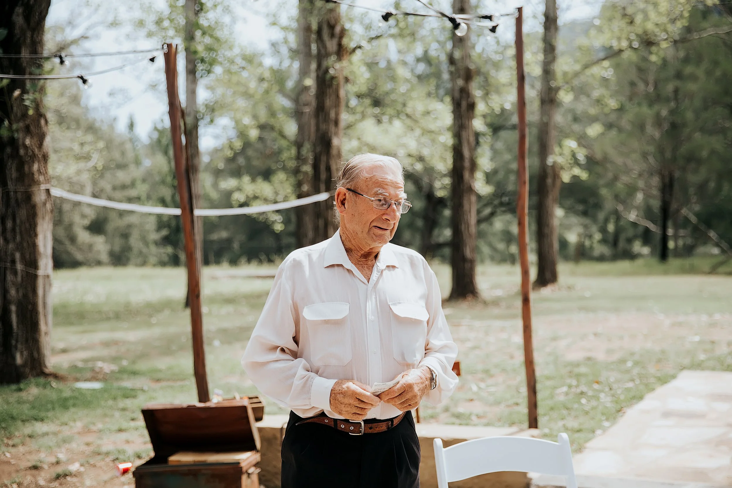 An elderly man with white hair and glasses stands outdoors among trees, wearing a white shirt and black pants, holding papers in his hands.