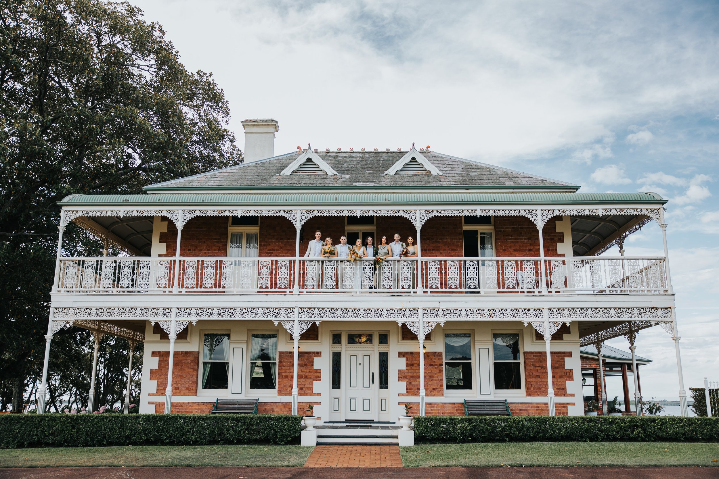 A large Victorian-style house with red brick walls, white lace trim on the porch, and a group of people dressed in formal or semi-formal attire standing on the upper balcony.