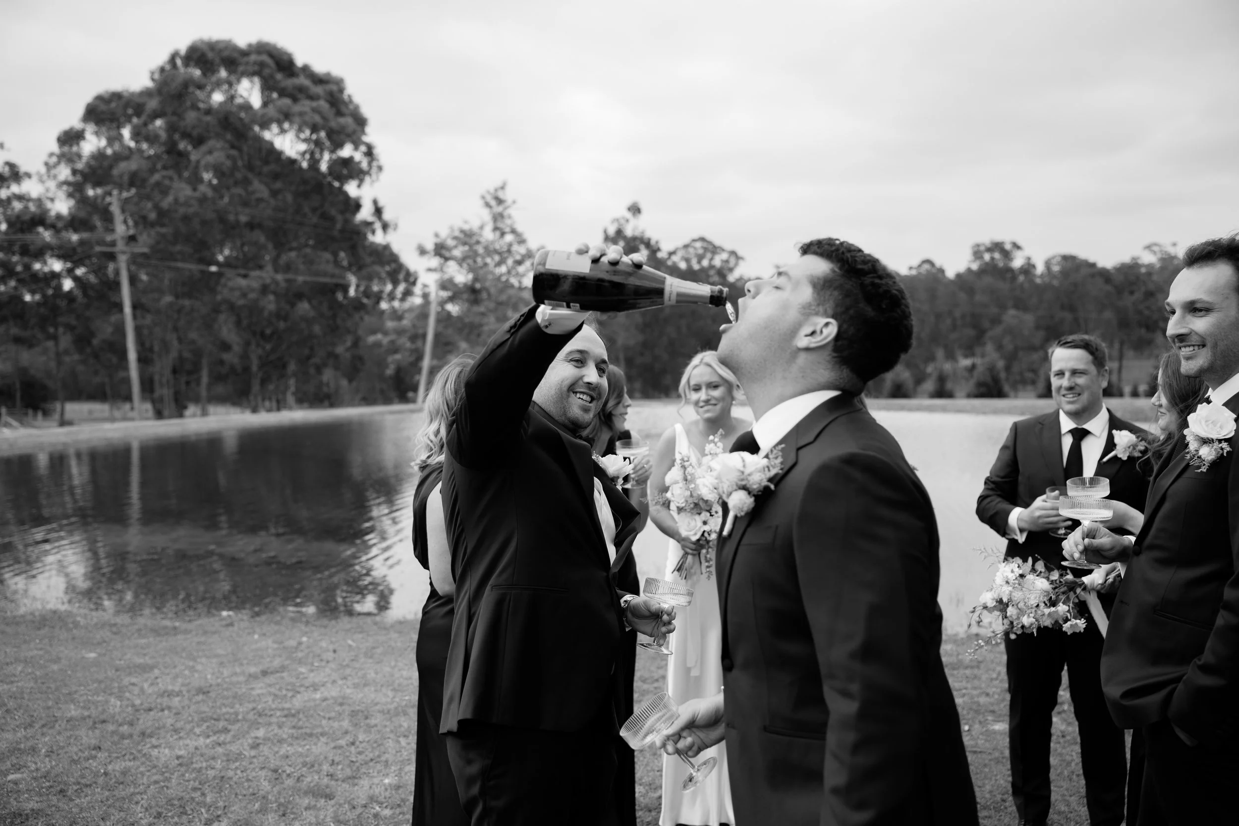 Black and white photo of a group of people at a wedding outdoors near a river, with trees in the background. One man is pouring champagne into a woman's mouth. The group is dressed in formal attire, holding glasses and flowers, and is smiling and enj