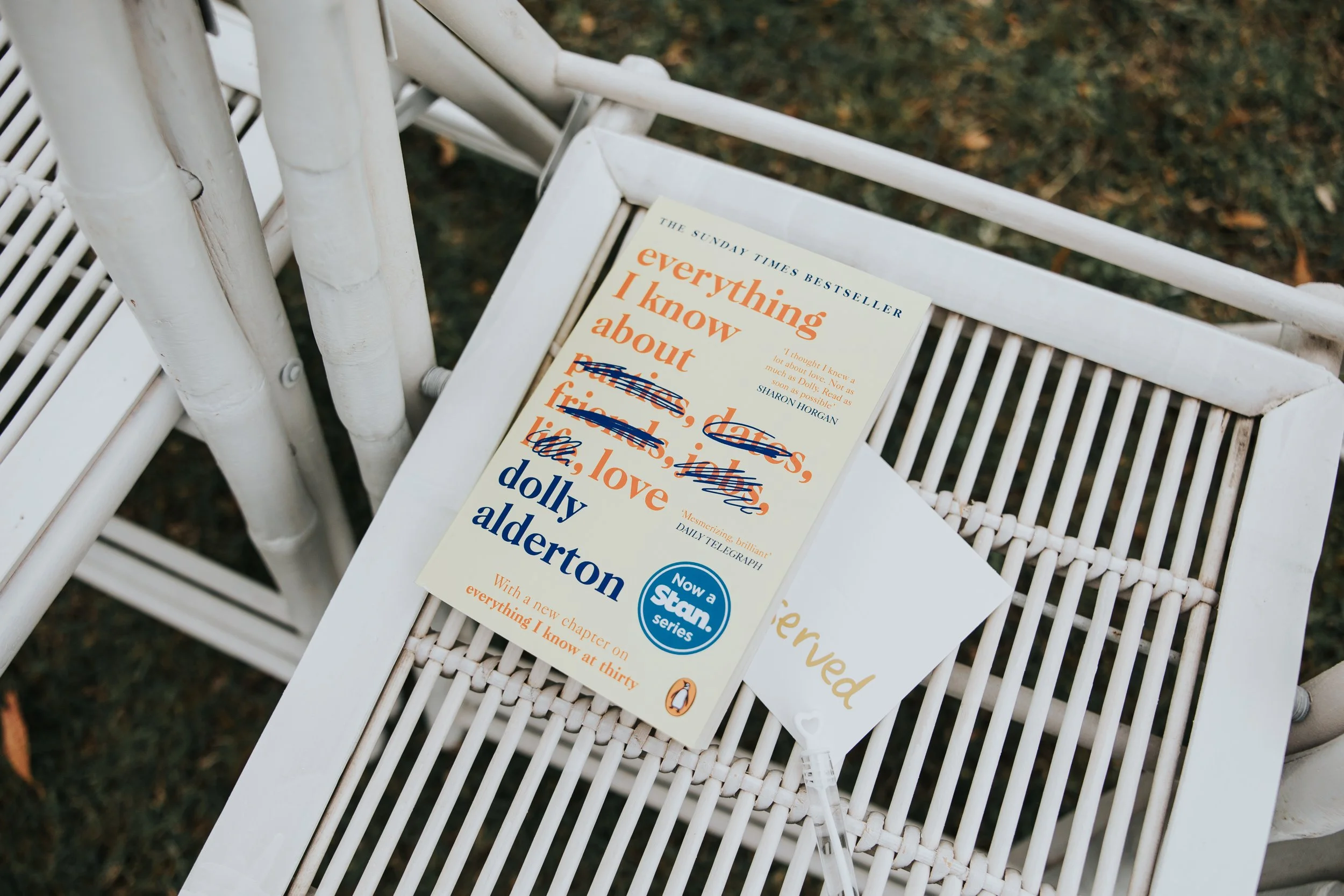 A white wicker chair outdoors with a paperback book titled "Everything I know about love" by Dolly Alderton resting on it. The background shows blurred foliage and grass.