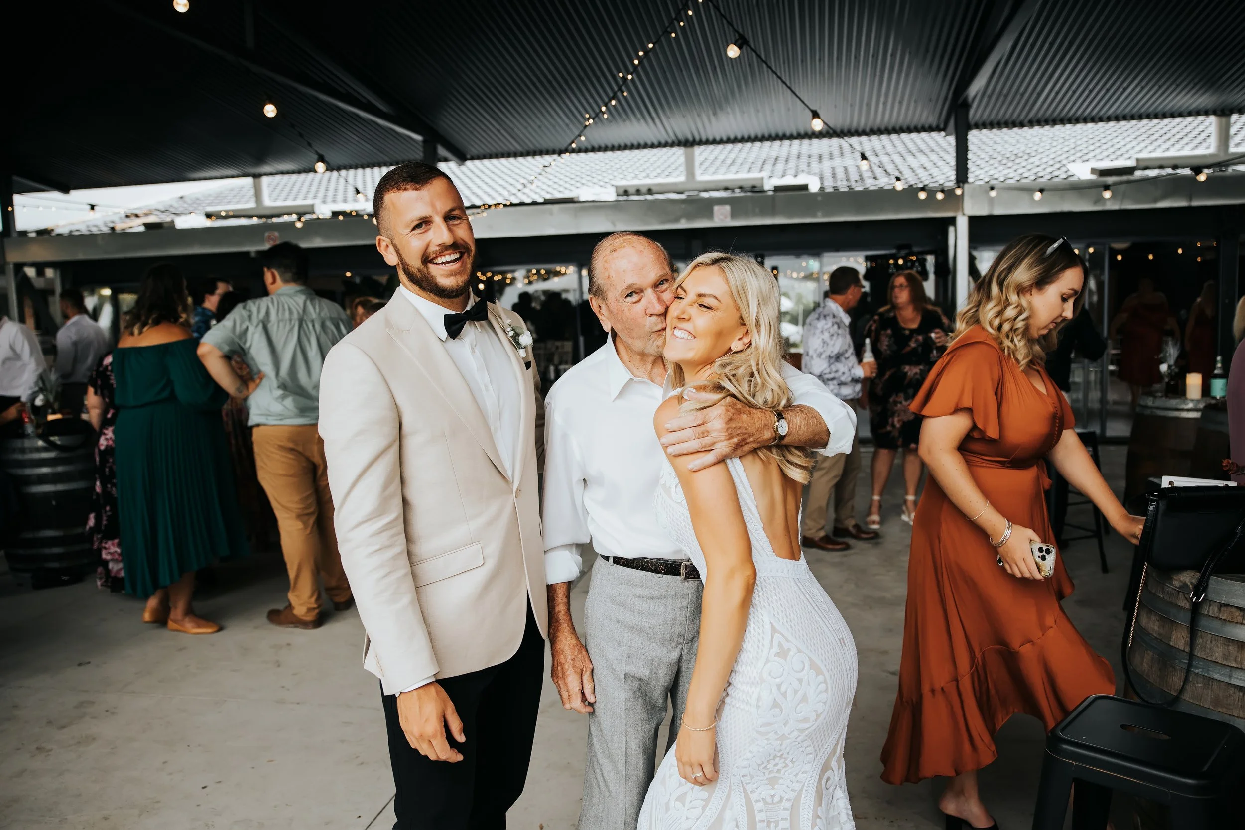 A group of people at a celebration, with a man in a tuxedo, a woman in a white dress, and an older man, hugging and smiling. Other guests are in the background, some looking at their phones, under a covered outdoor area with string lights.