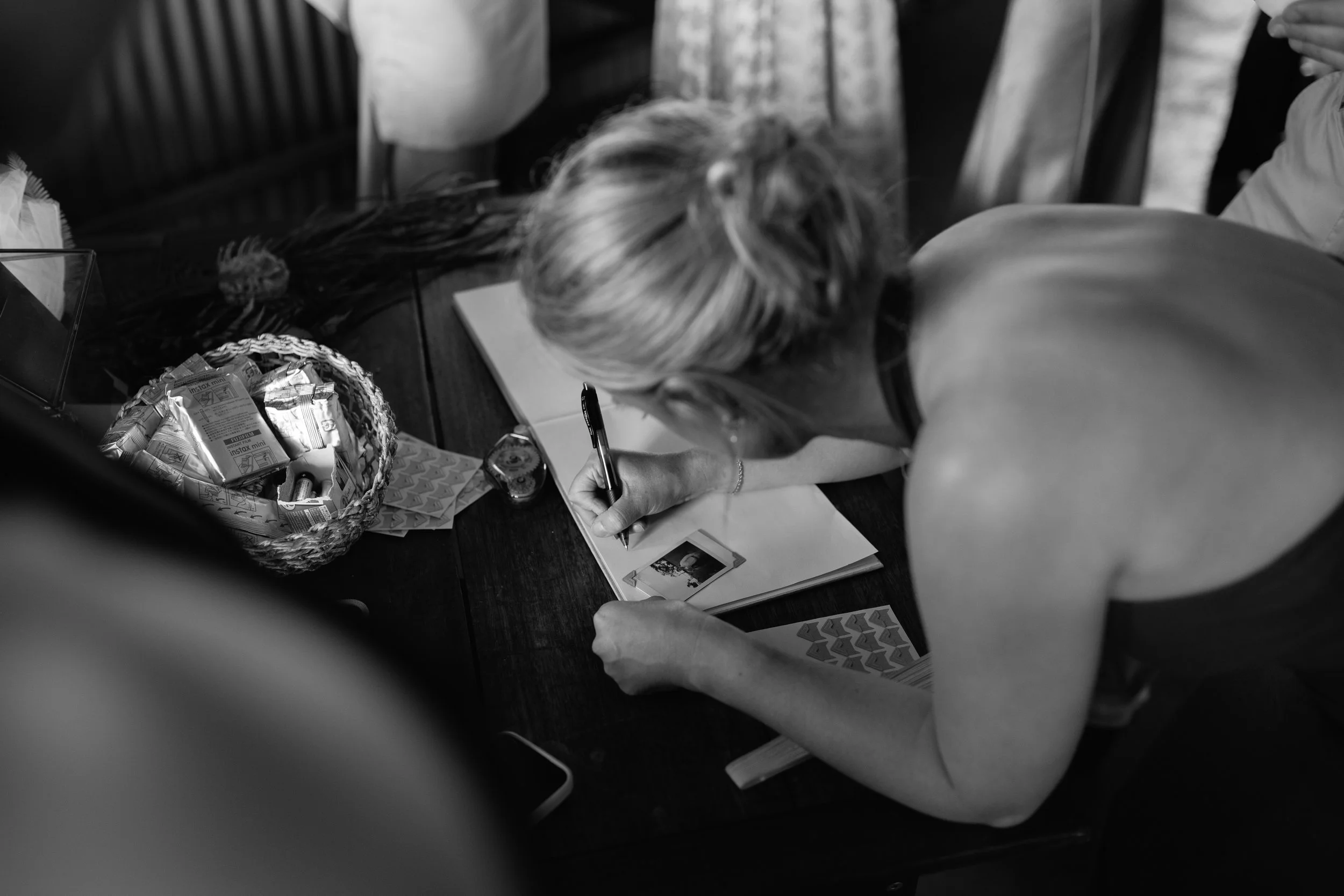 People gathered around a table, one person writing in a notebook, with a basket of snacks and various papers on the table.
