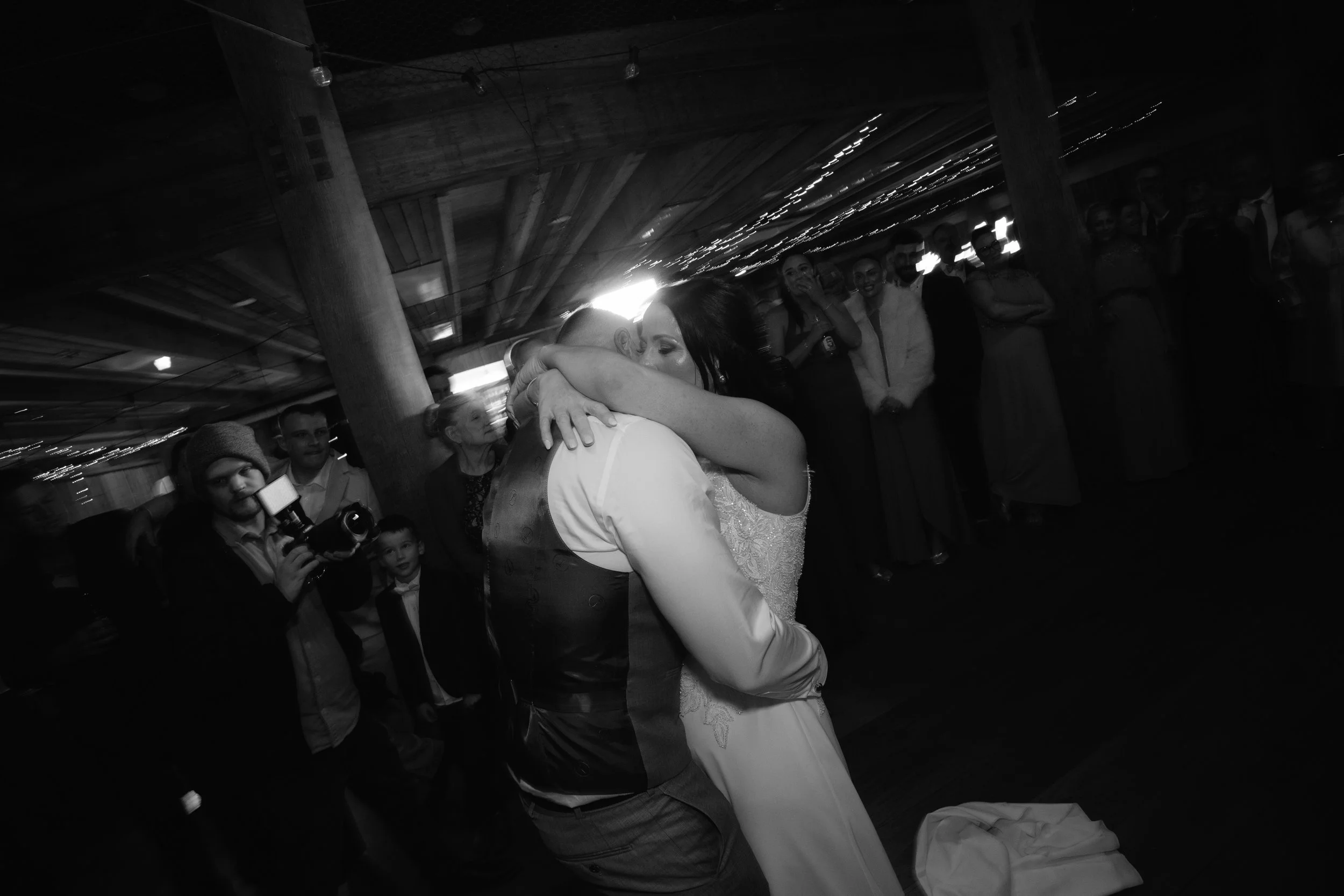A couple dancing closely at a social gathering or party, with several onlookers and photographers watching, in a dimly lit indoor setting.