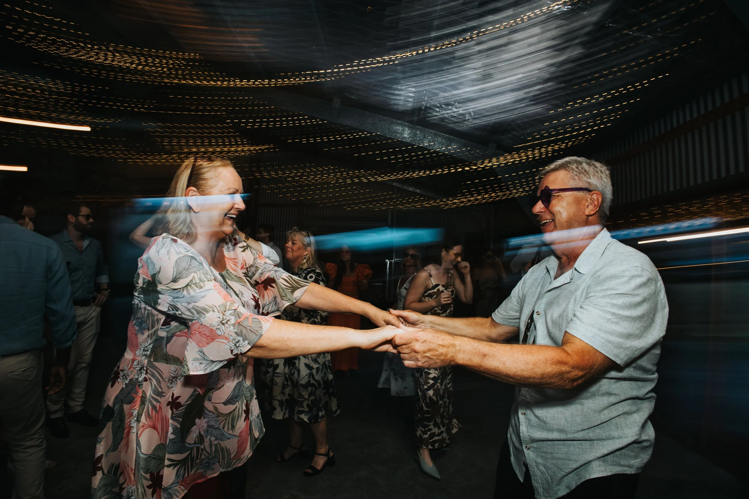 Two people, an older man wearing sunglasses and an older woman in a floral shirt, are dancing together while holding hands at a lively indoor party with other guests in the background and decorative ceiling lights.