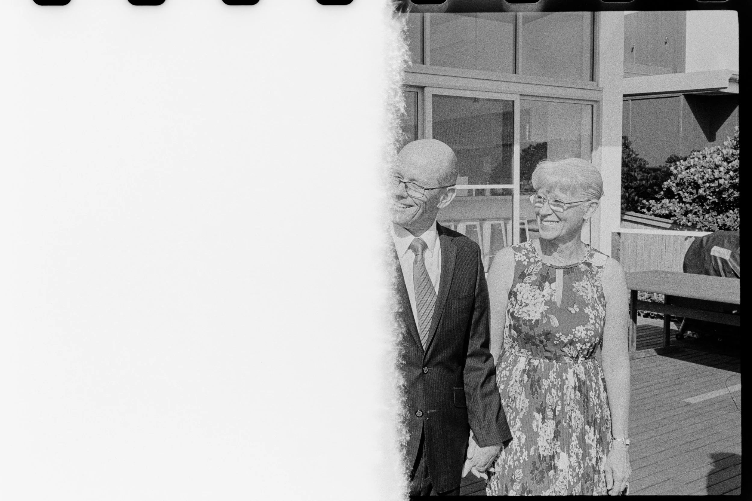 A black-and-white photo of a man in a suit and a woman in a floral dress, both smiling, standing outdoors on a wooden deck in front of large windows, with trees and a building in the background.