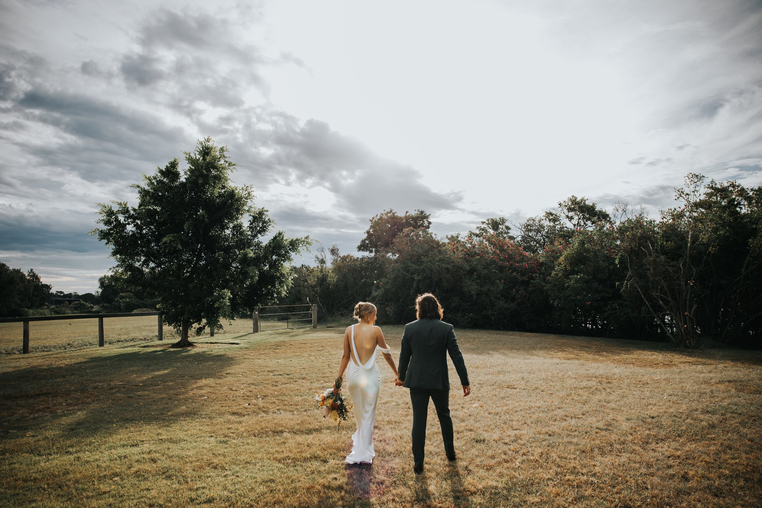 A man and woman walking hand in hand outdoors on a grassy field, with trees and a cloudy sky in the background. The woman is wearing a white dress, and the man is in a dark suit.