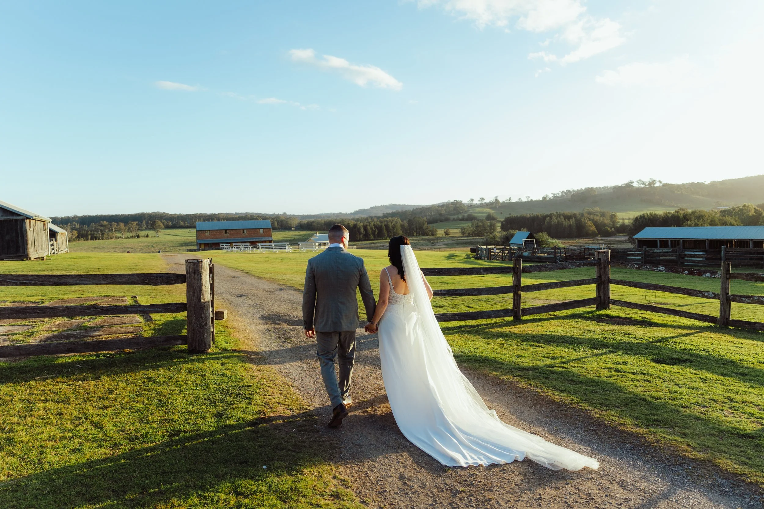 A bride and groom walking hand in hand in a rural farm setting with wooden barns, green fields, and a distant hill, during sunset.