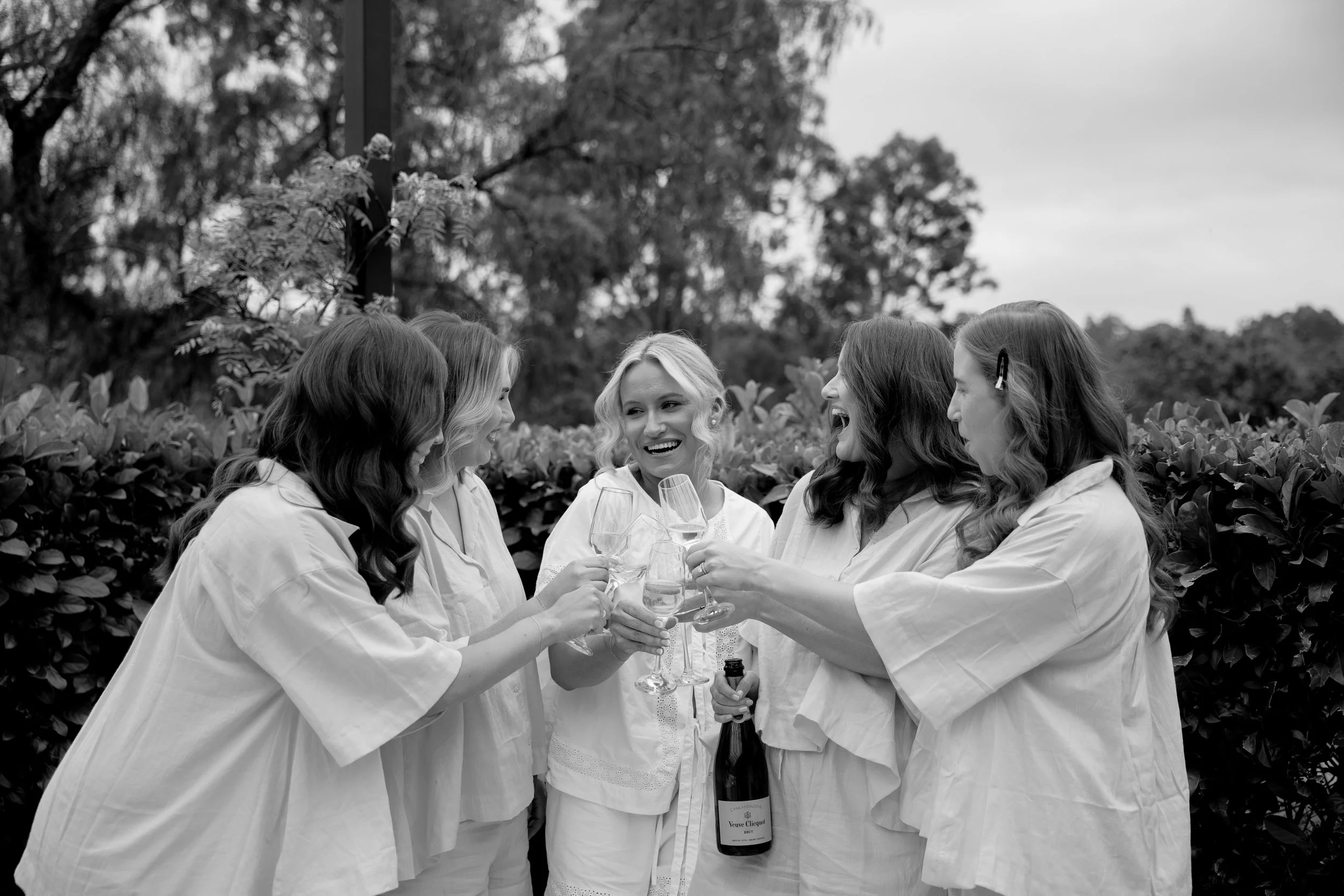 Group of five women in white dresses celebrating with champagne outdoors, one woman holding a bottle of Veuve Clicquot champagne, smiling and toasting.