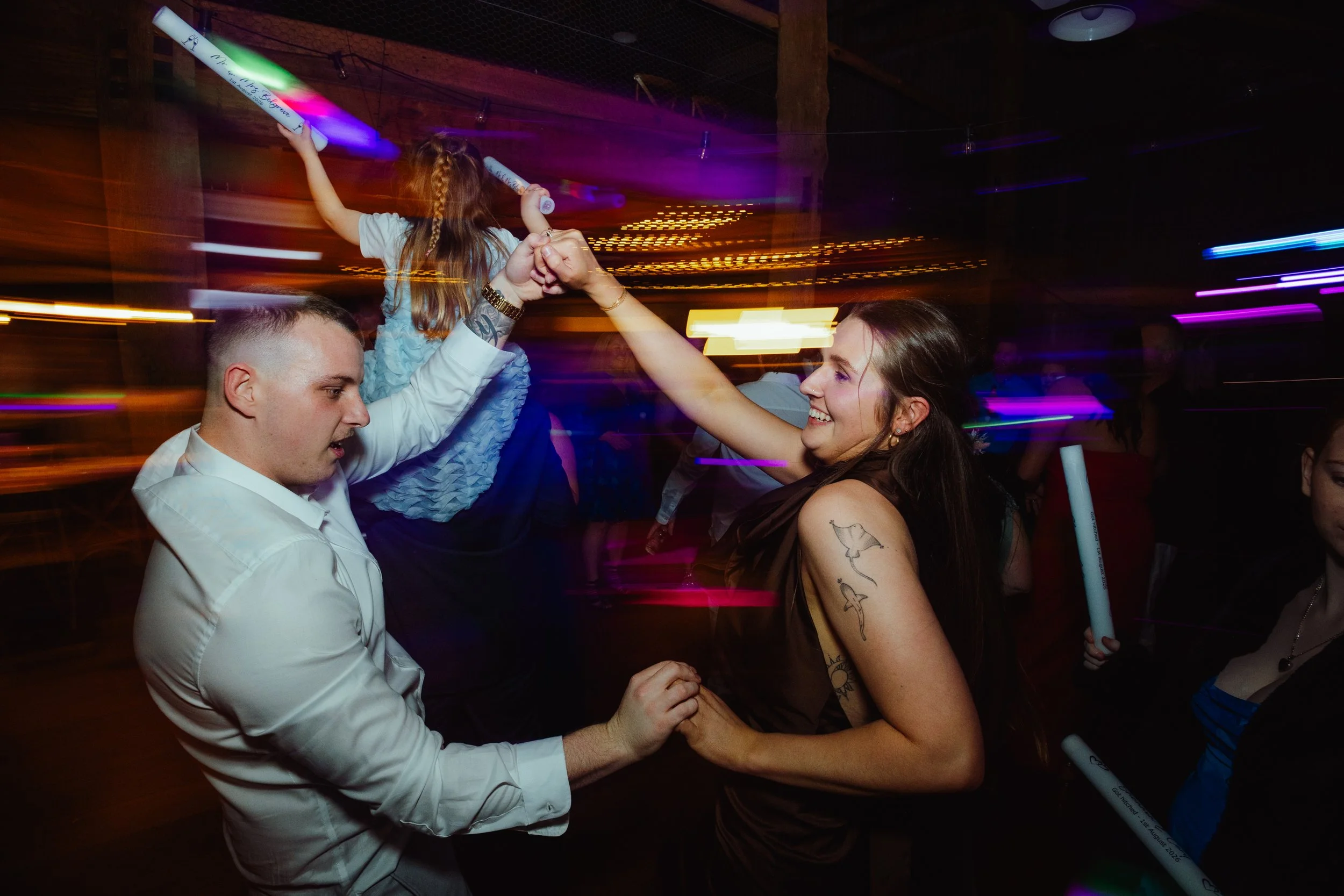 A man and a woman are dancing and holding hands at a party, with children and colorful lights around them.