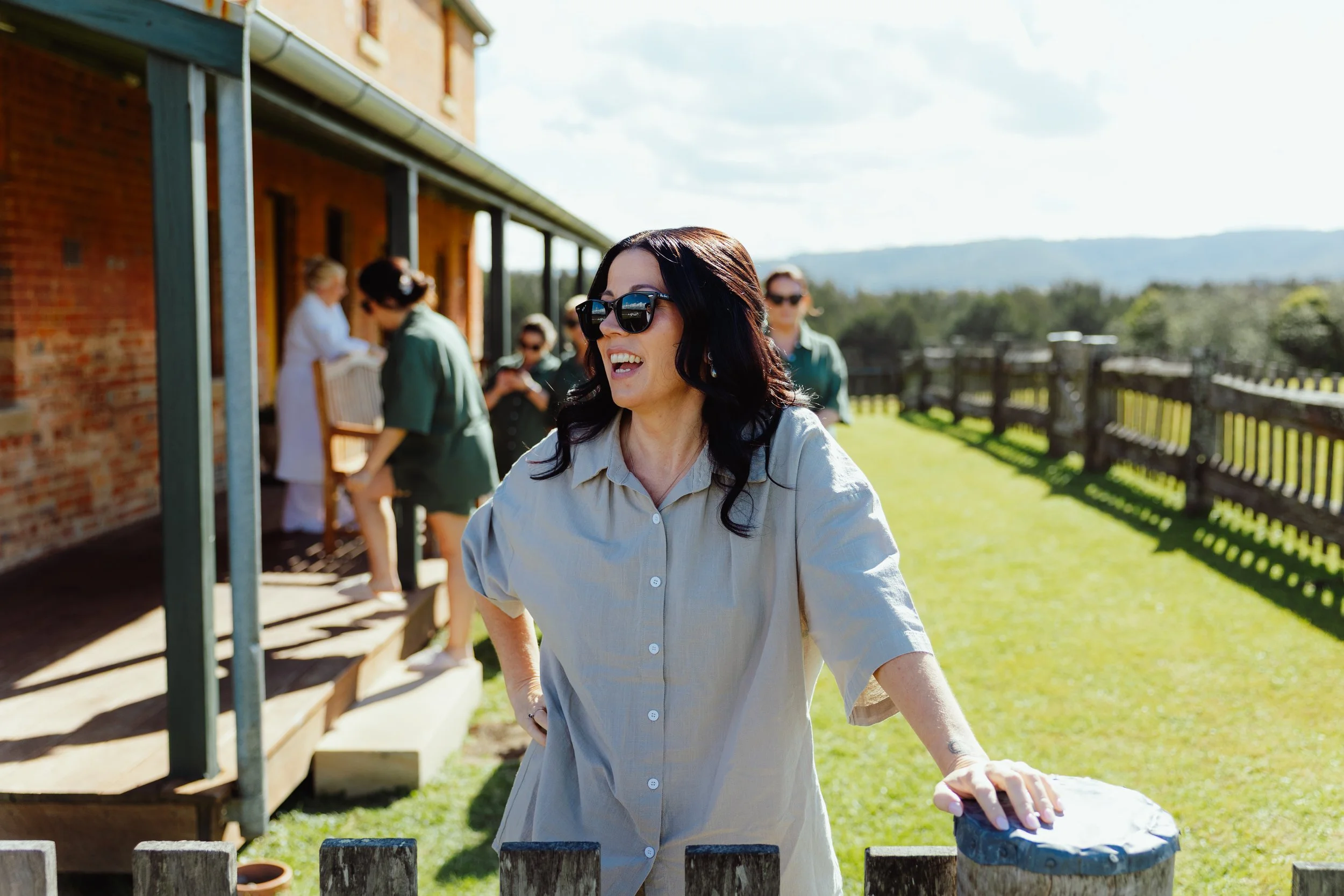 A woman with dark hair, wearing sunglasses and a light-colored shirt, smiling outside on a sunny day with a group of women in the background near a brick house and a wooden fence.