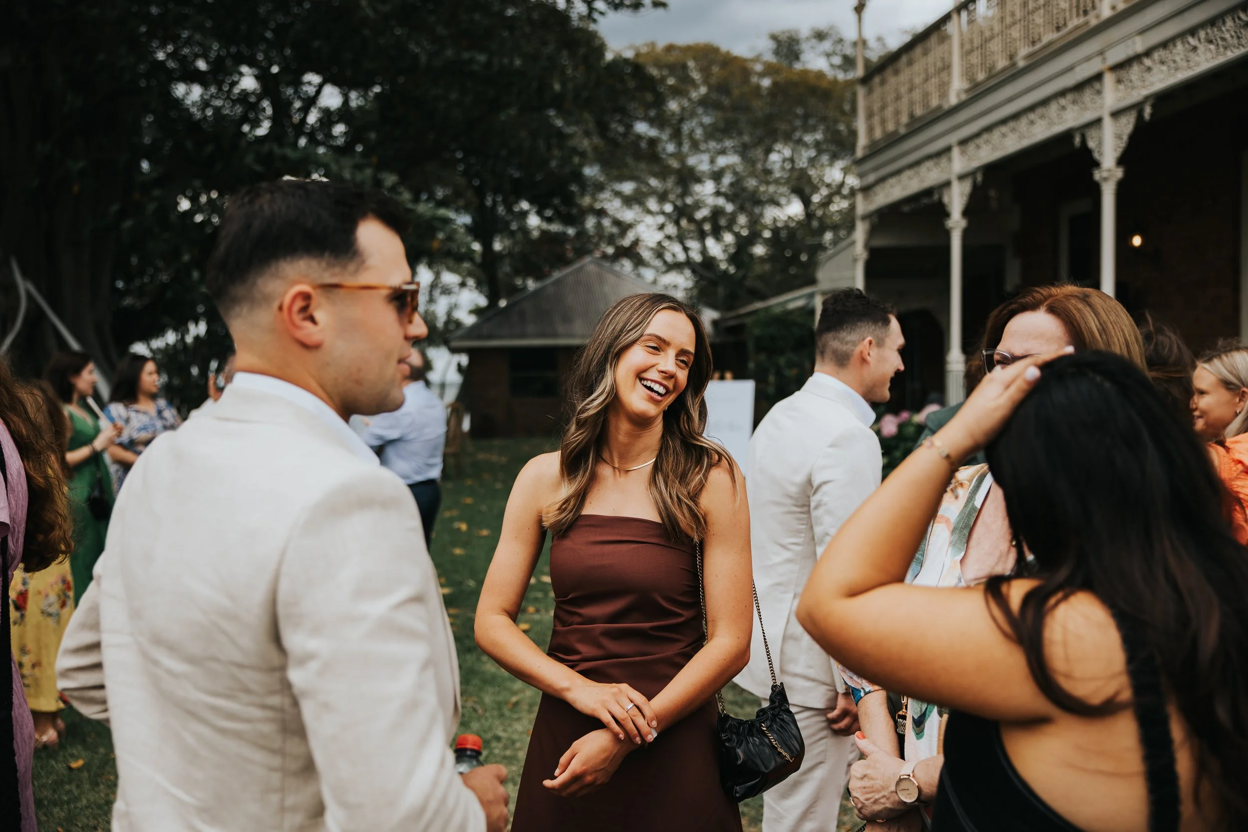 Group of people gathered outdoors, socializing at a formal event, with a woman smiling in the center, wearing a brown strapless dress.