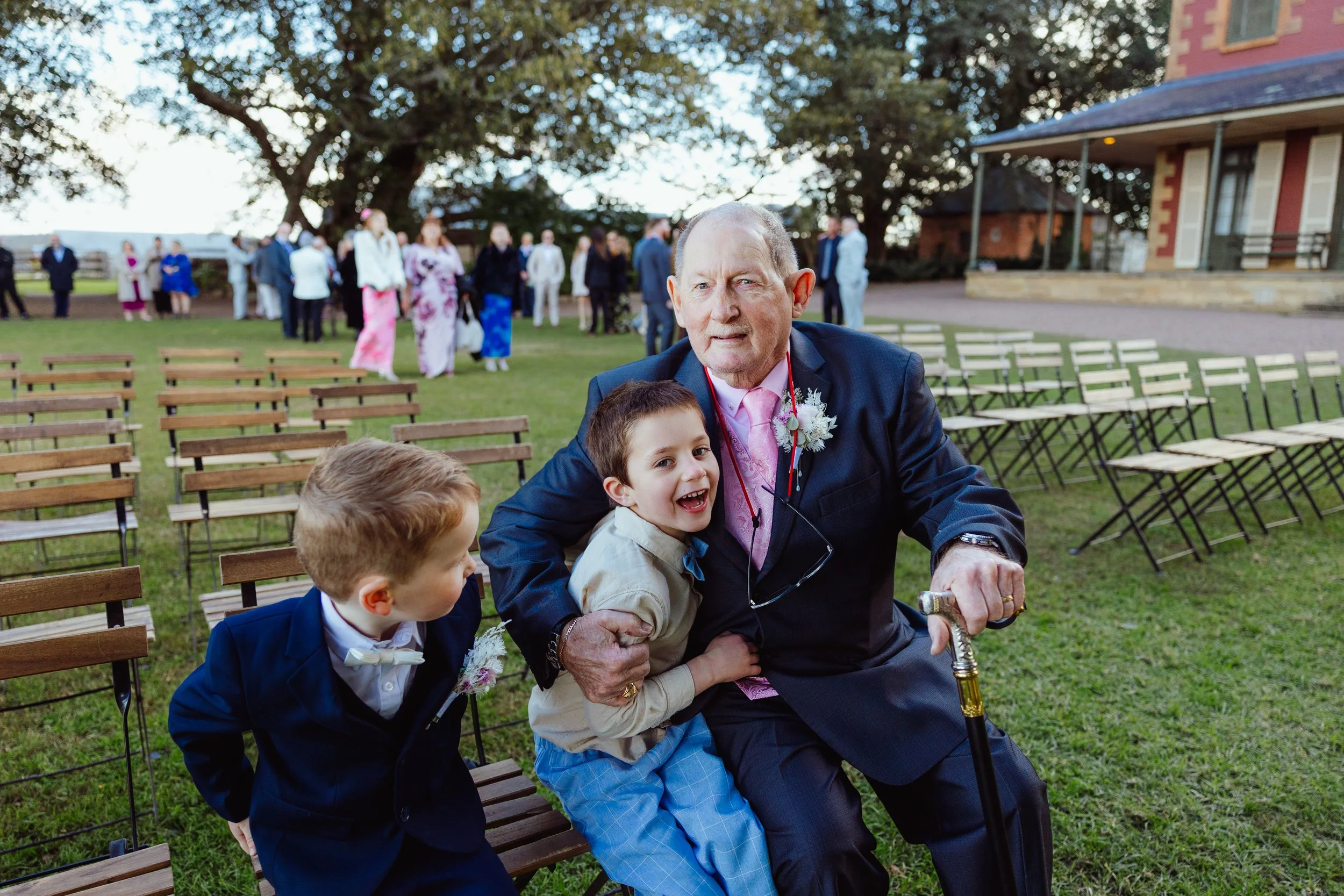 A man with a cane and children sitting on a wooden bench at an outdoor event, with a group of people in traditional attire gathered behind them under large trees.
