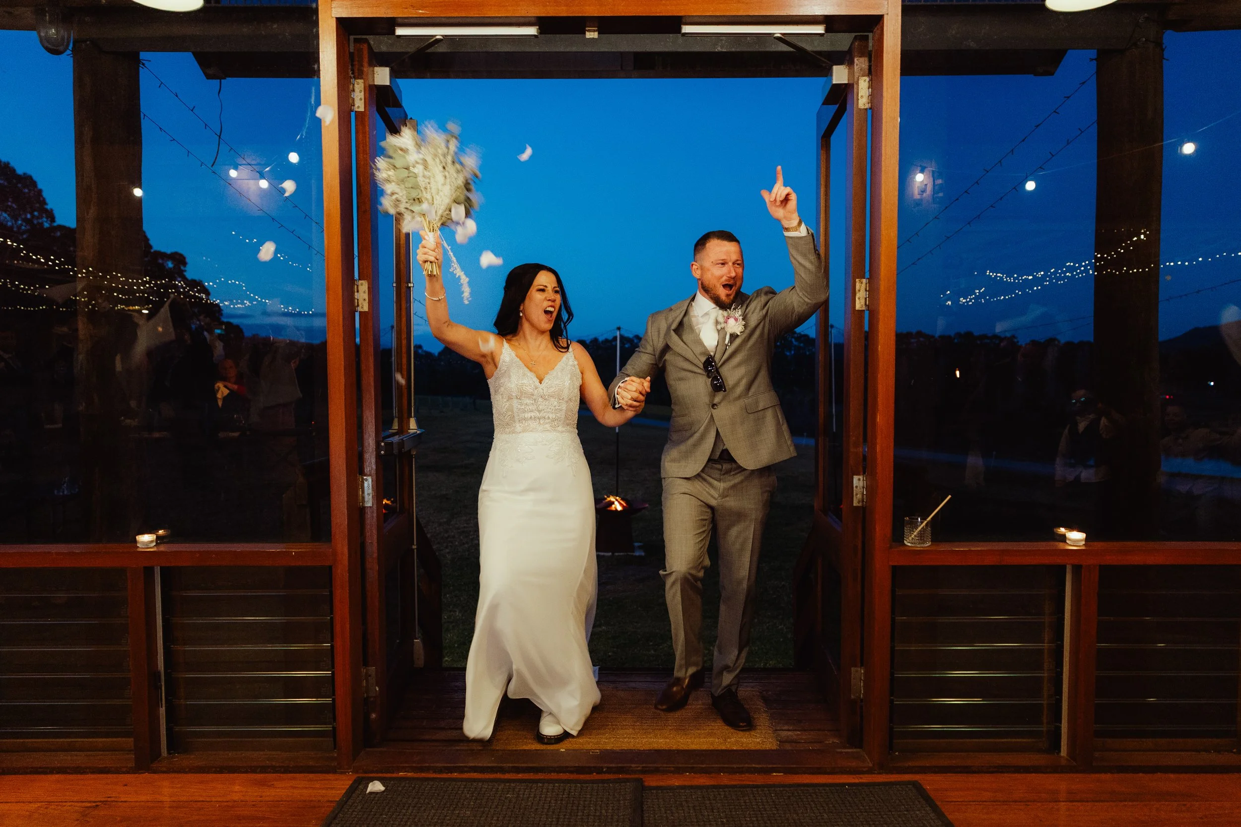 A bride and groom celebrating as they walk through a wooden doorway at dusk, with the bride holding a bouquet and the groom raising his finger in the air. Confetti is falling around them, and string lights are visible in the background.