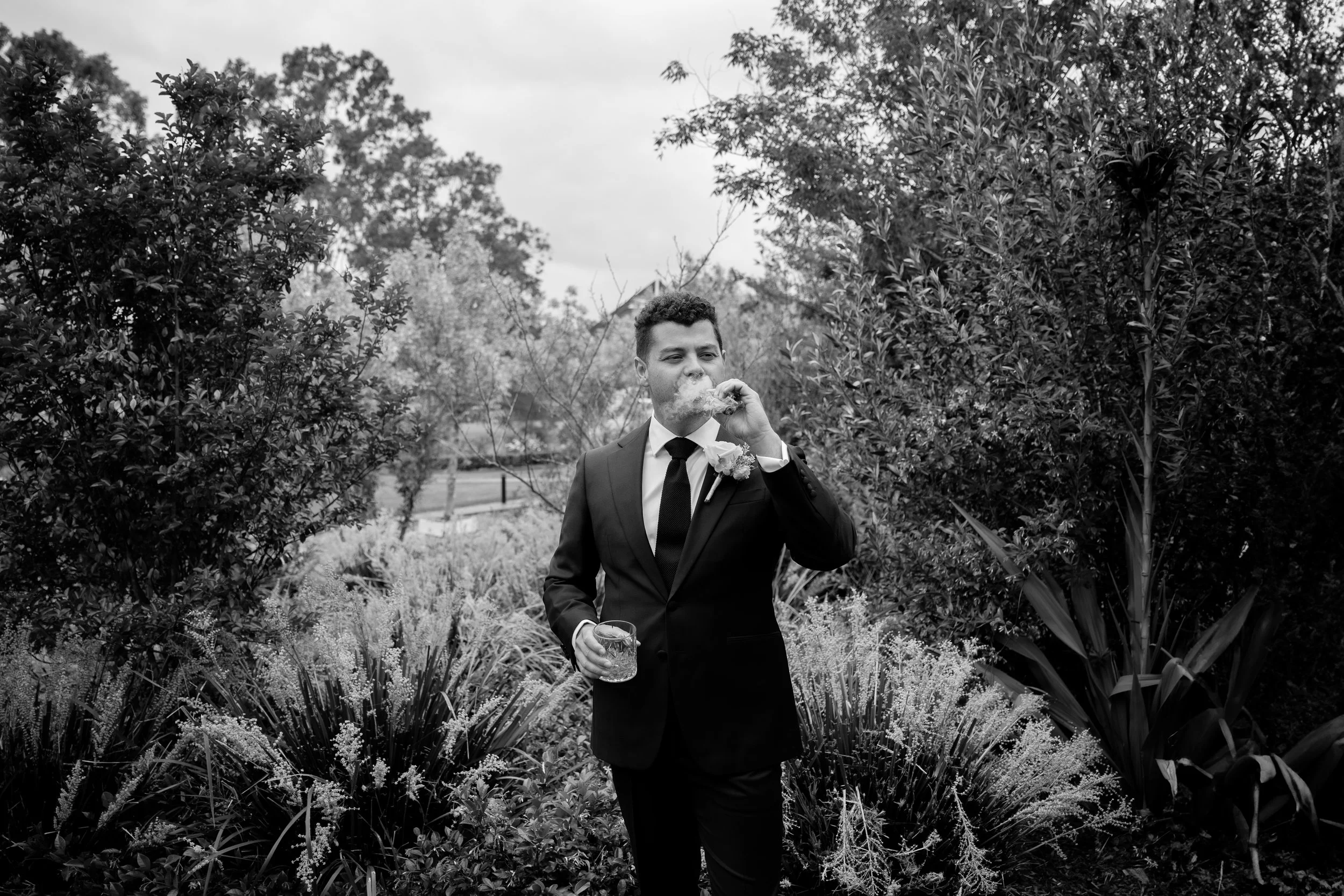 A man in a suit, possibly a groom, standing outdoors among plants and trees, smoking and holding a drink in a garden or park setting.