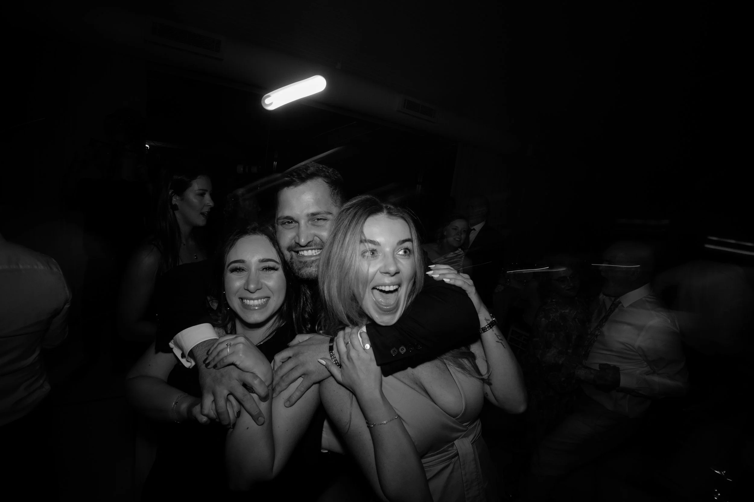Group of people smiling and hugging at a social event or party, indoors with a dark background and bright overhead lighting.