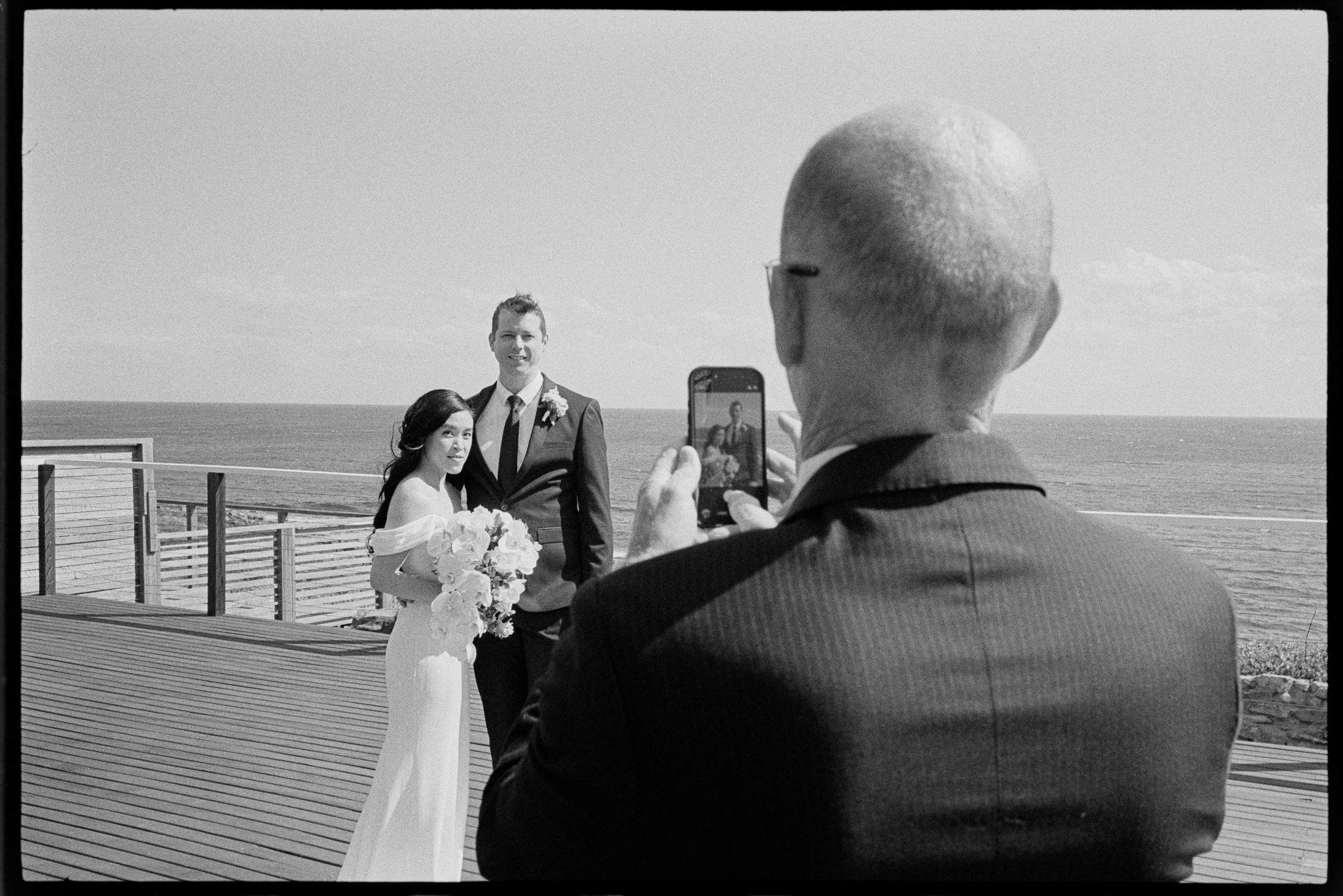 A couple getting married outdoors by the ocean, with a man in a suit taking a photo of them on a smartphone, during a sunny day.