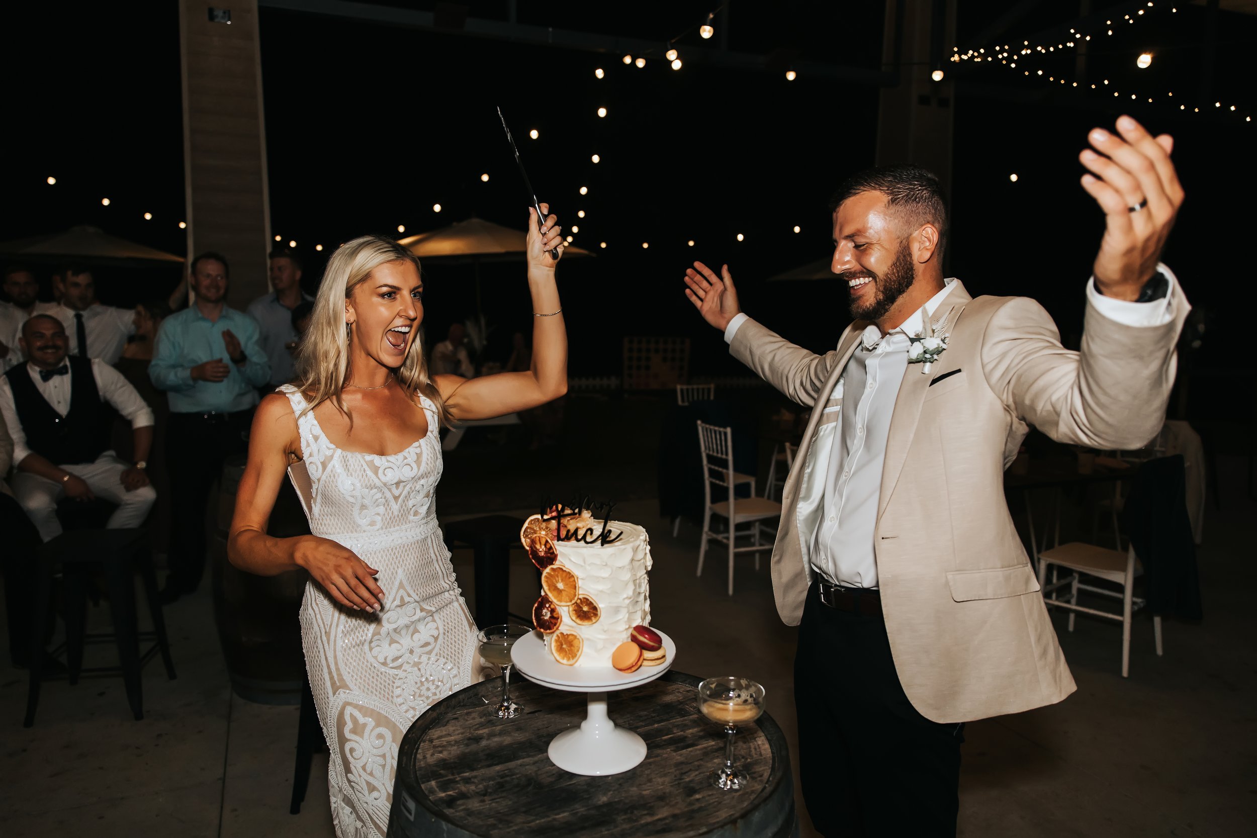 A bride and groom celebrating at their wedding reception, with the bride holding a cake knife preparing to cut a cake decorated with dried fruit, while guests clap and smile in the background under string lights.