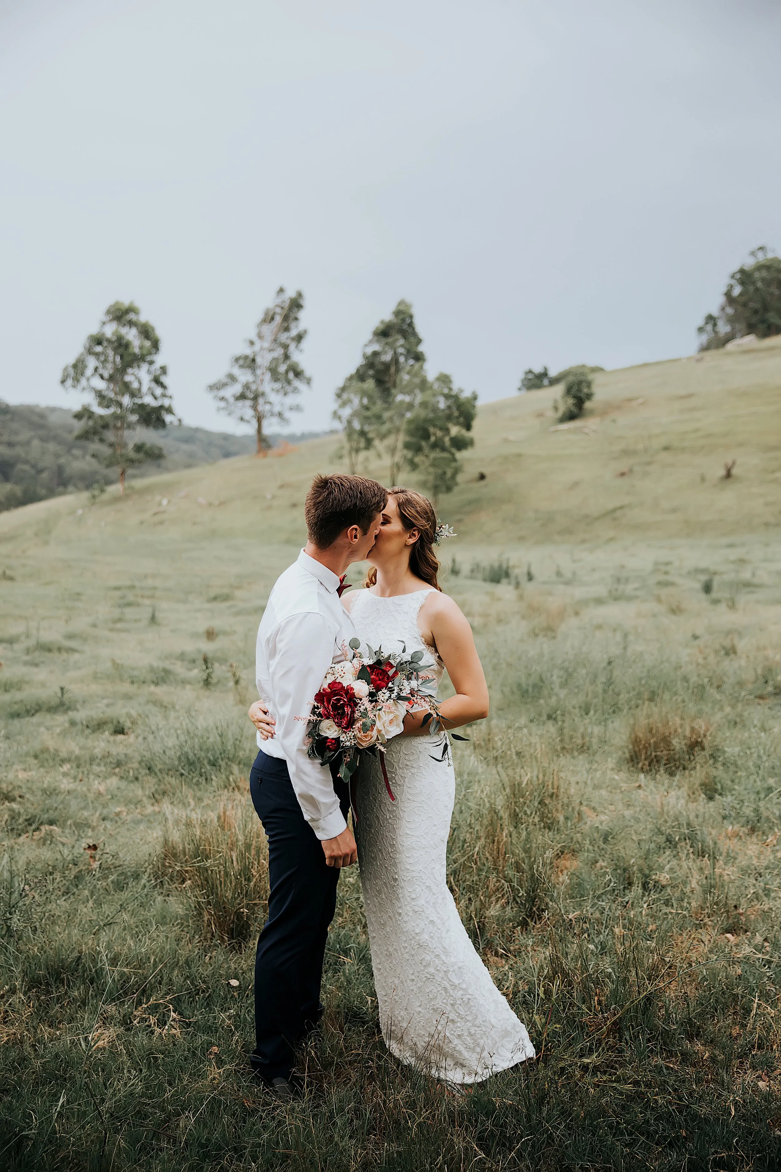 A bride and groom sharing a kiss in a grassy outdoor field with rolling hills and scattered trees in the background.