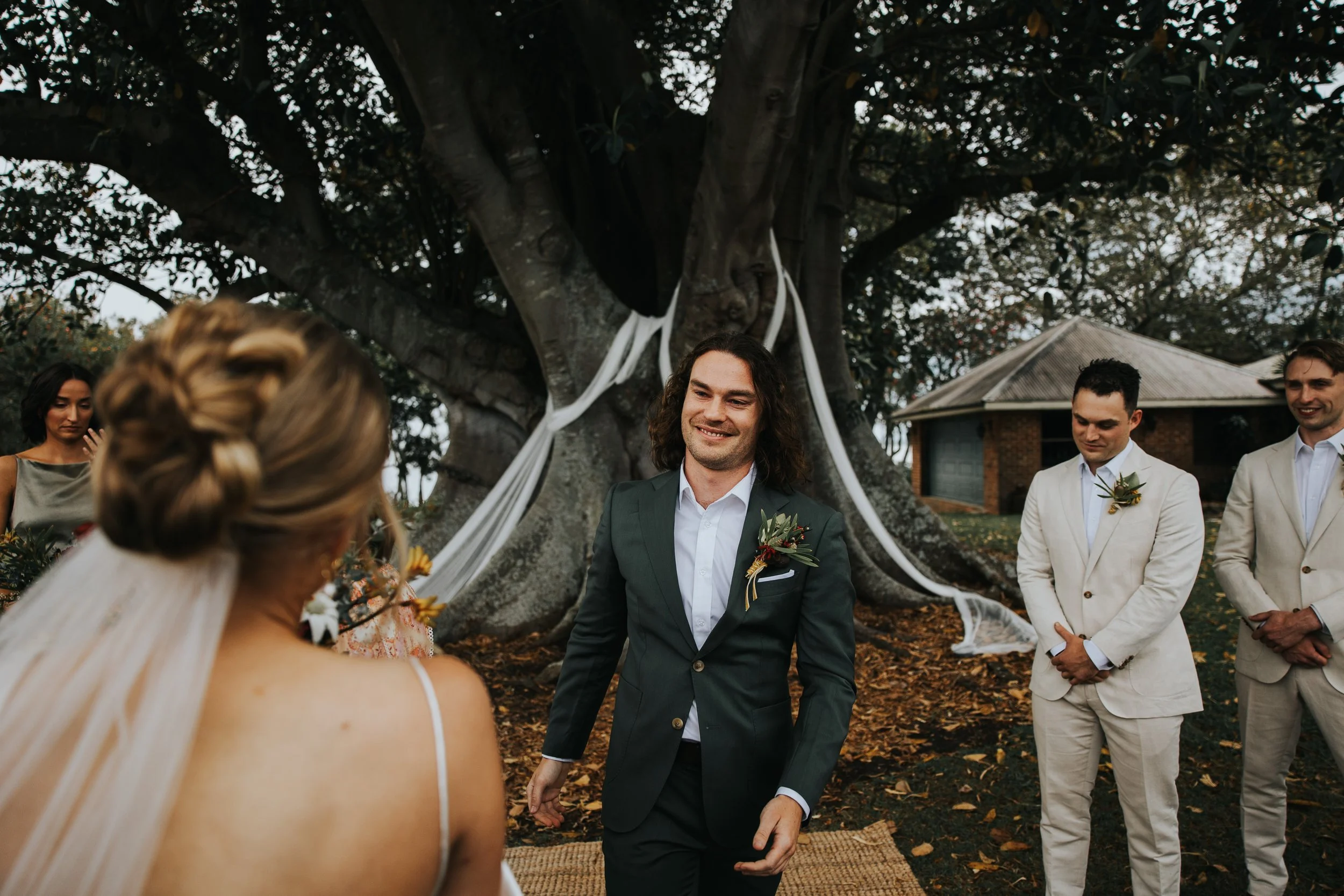 A wedding ceremony outdoors with a groom holding hands with a bride, standing under a large tree decorated with white fabric. Bridesmaids and groomsmen look on, dressed in formal attire, in front of a house with a slanted roof. The scene is set in a 
