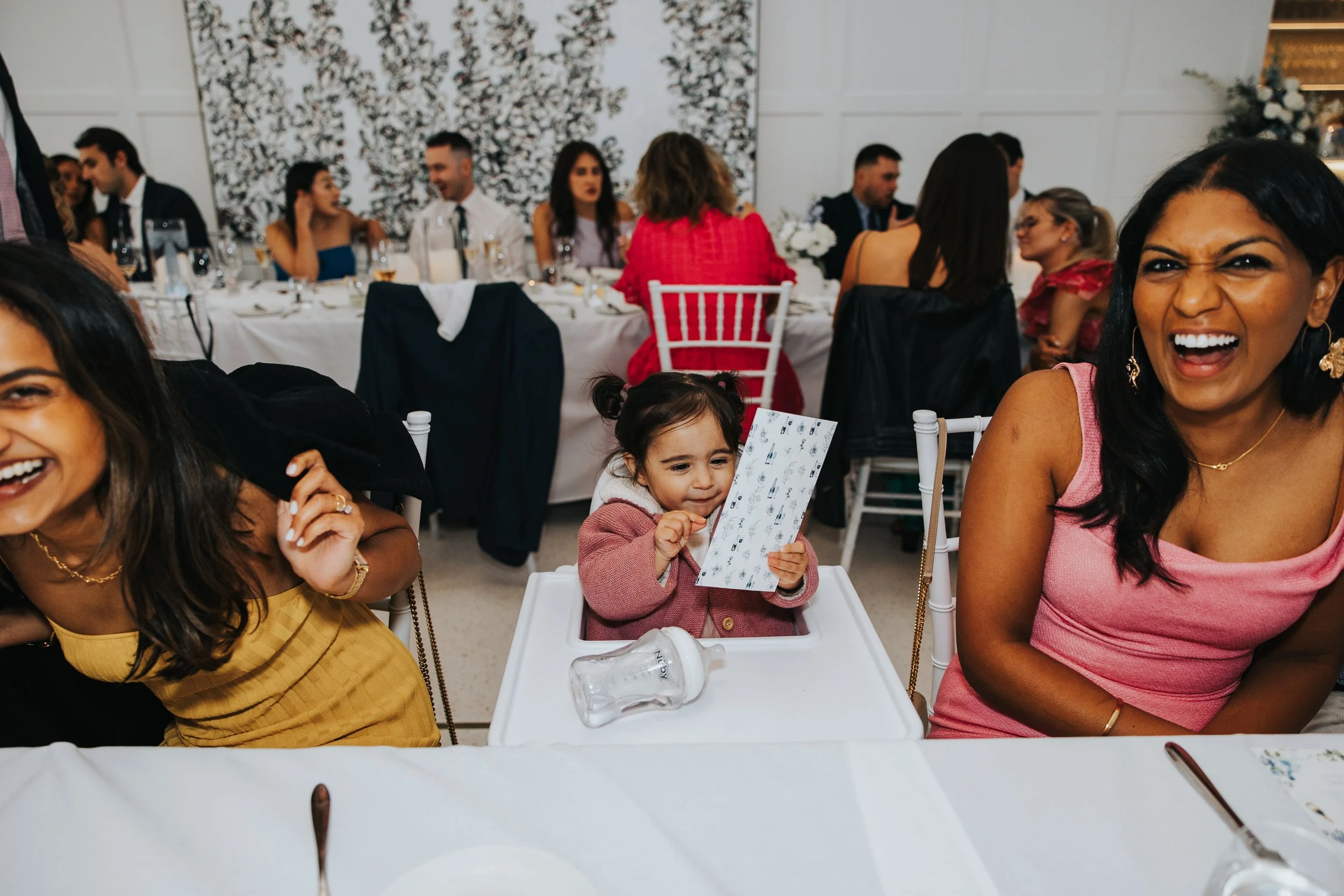 People gathered around a dining table at a celebration or event, engaging in conversations, with some smiling and laughing, in a decorated indoor setting.