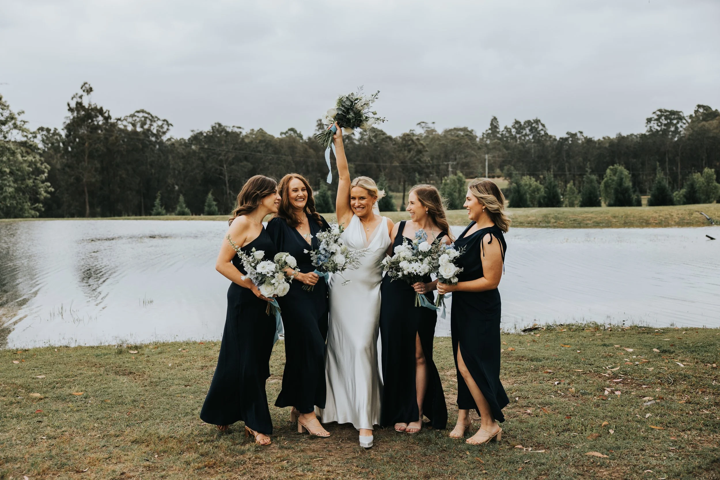 A group of five women standing outdoors by a lake, with trees in the background. The woman in the center, dressed in white, is holding her bouquet high, surrounded by four women in black dresses, each holding bouquets, celebrating at a wedding event.