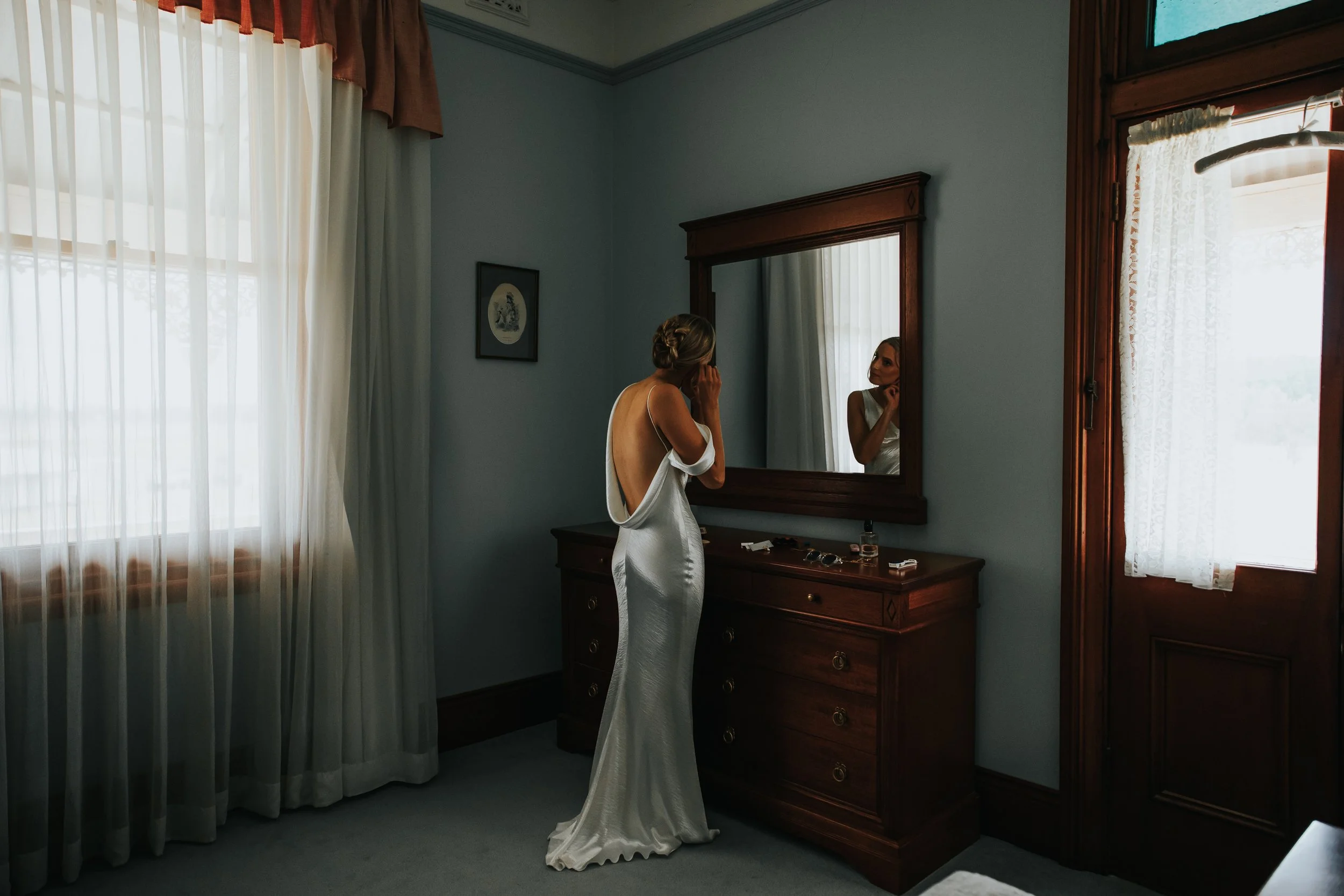 A woman in a white satin dress looking into a mirror in a bedroom, with natural light coming through the window with sheer curtains.