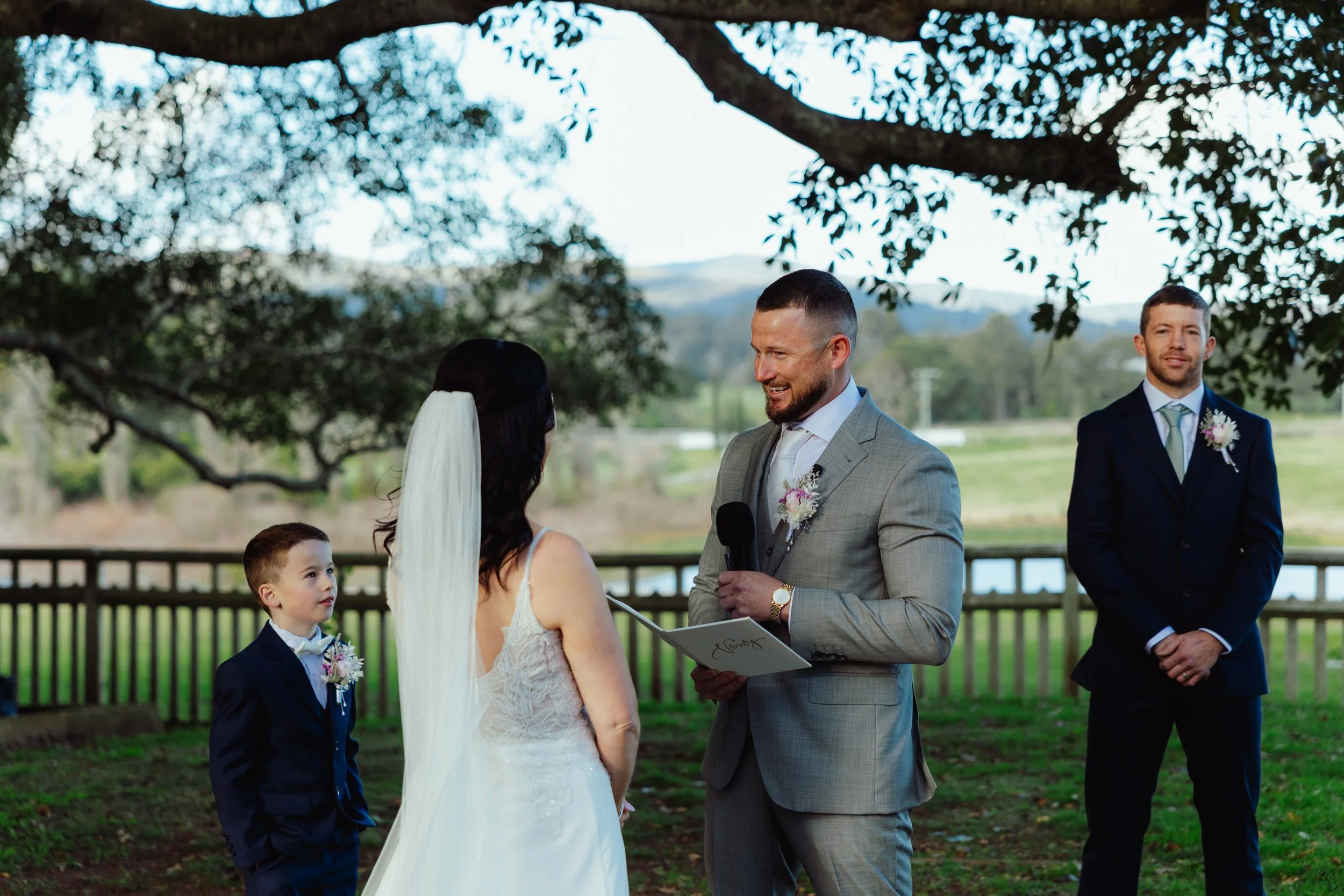 A wedding ceremony taking place outdoors under a large tree, with a man in a gray suit holding a microphone, a woman in a wedding dress, a young boy in a suit, and another man in a dark suit with a boutonniere. The scene is set in a natural environme