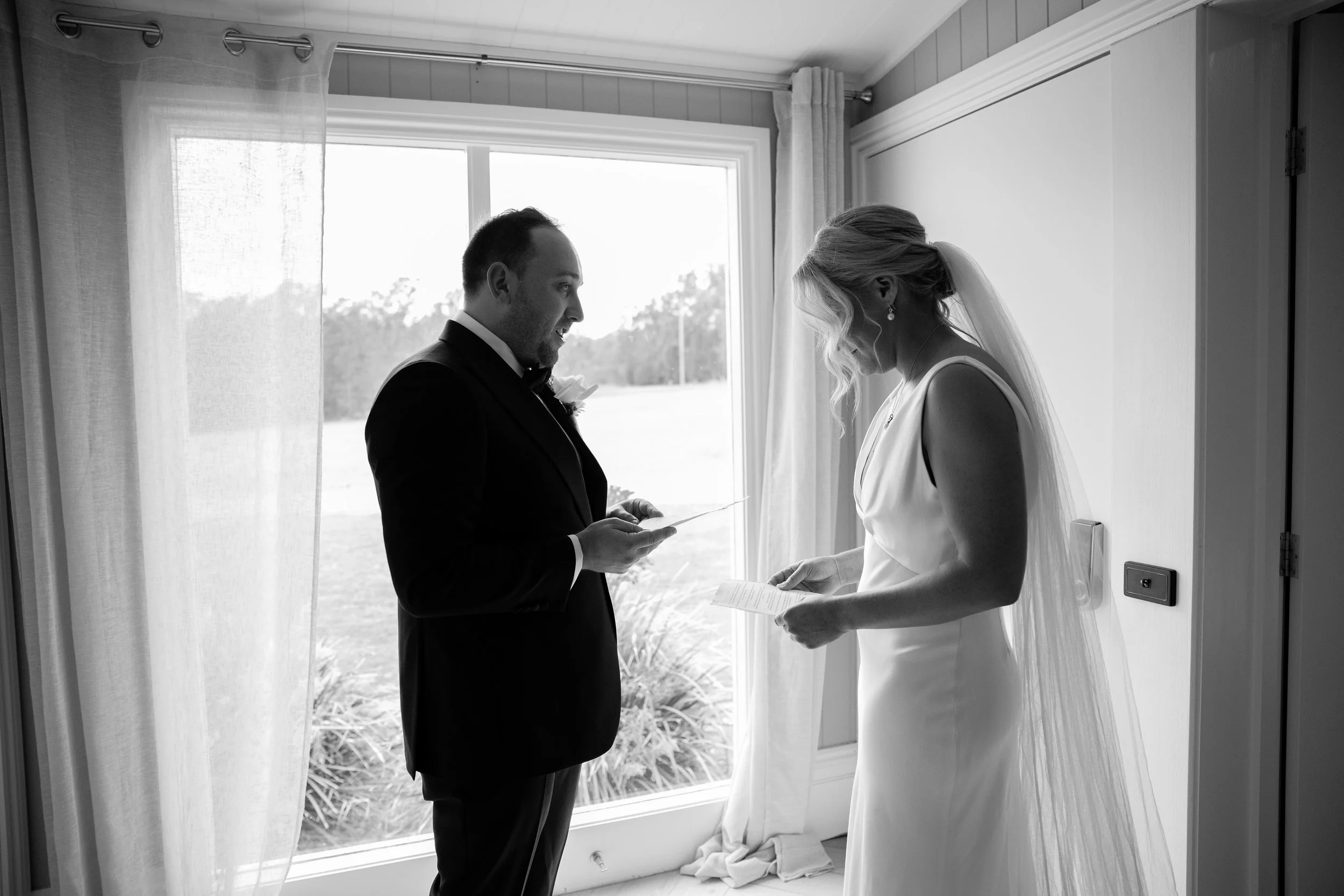 A bride and groom standing by a window, facing each other during their wedding ceremony, with the groom holding a small piece of paper or card.