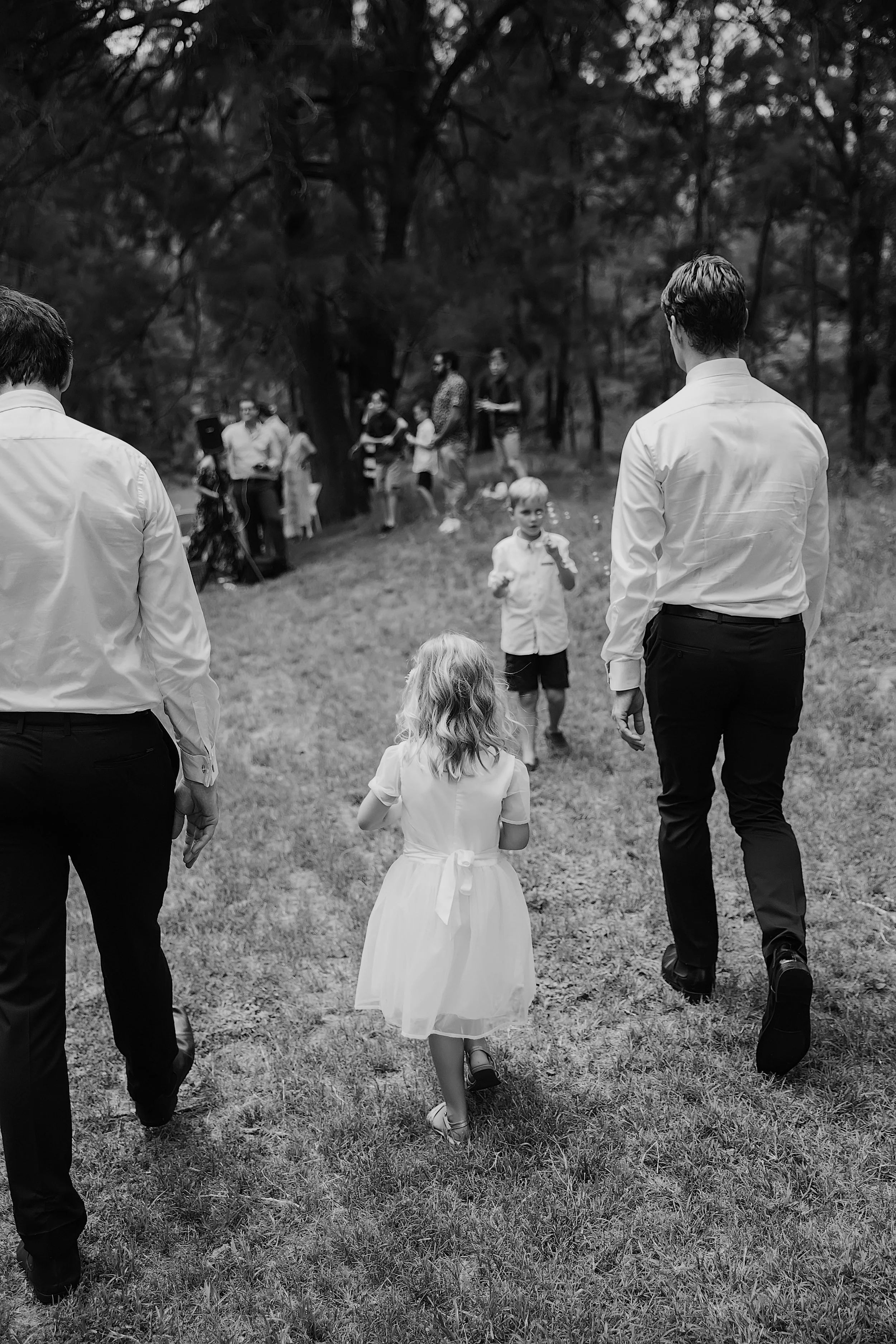 Black and white photograph of a group of people walking outdoors in a grassy area surrounded by trees. In the foreground, a young girl in a white dress walks ahead, with two men in white shirts on either side of her. In the background, there are more