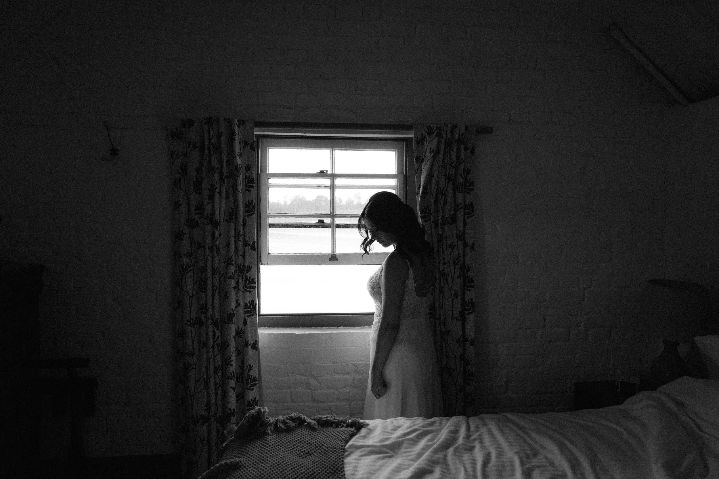 A woman in a white dress stands in front of a window, looking down, in a dimly lit room.