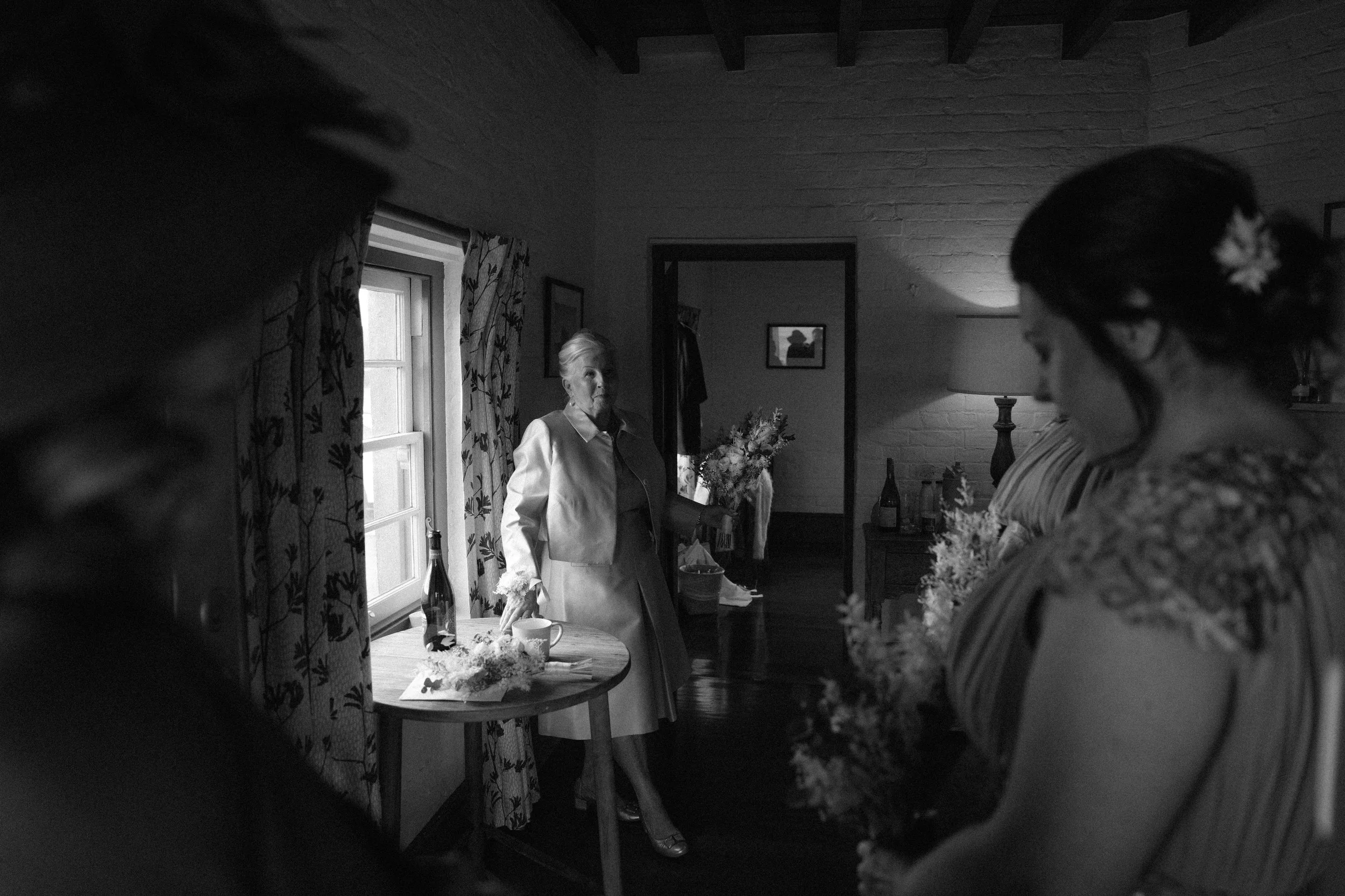 A black and white photo of a woman in a dress standing near a table with flowers and beverages, with another woman with flowers in her hair in the foreground.