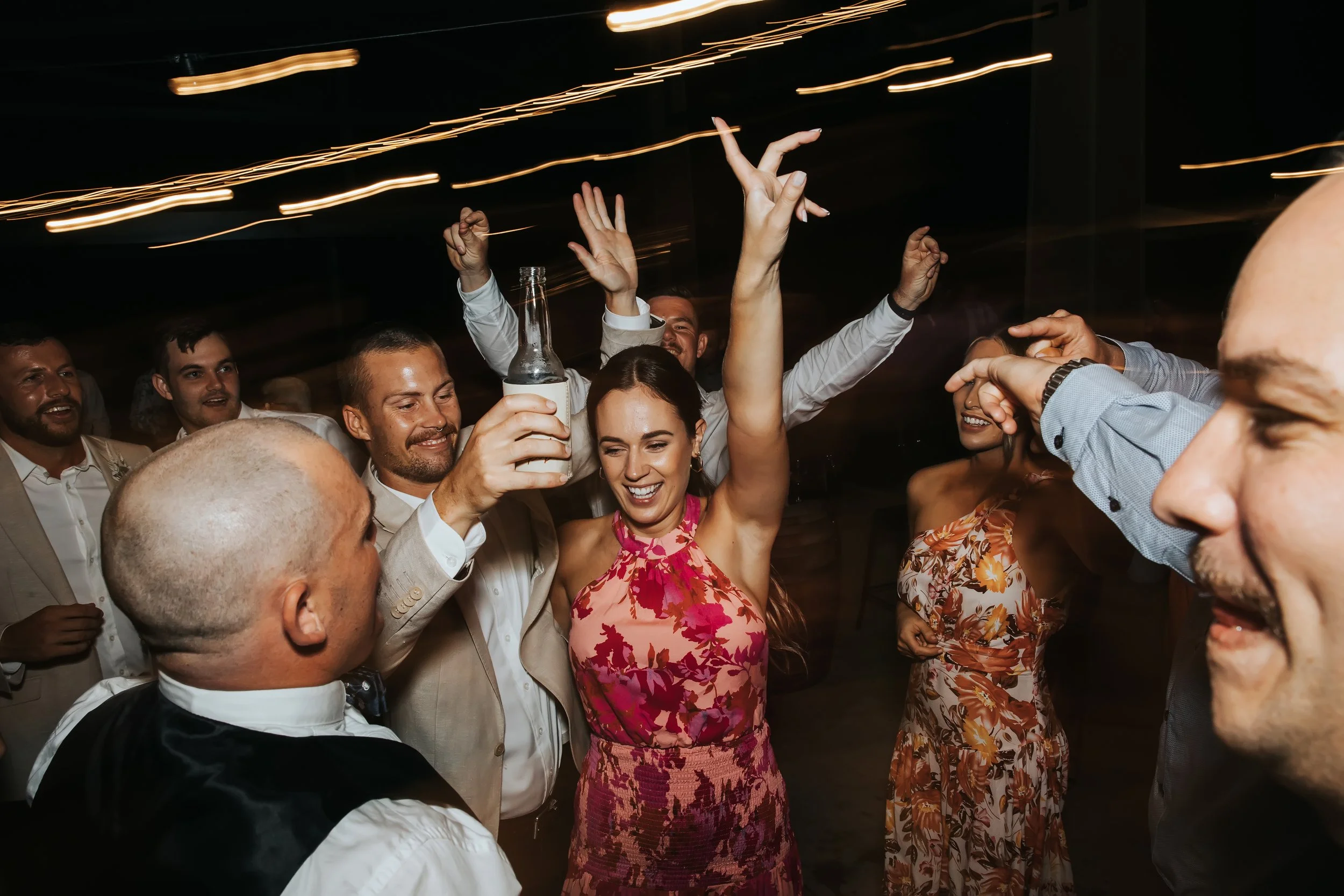 People dancing and celebrating at a party, some holding drinks, with colorful lights and smiling faces.
