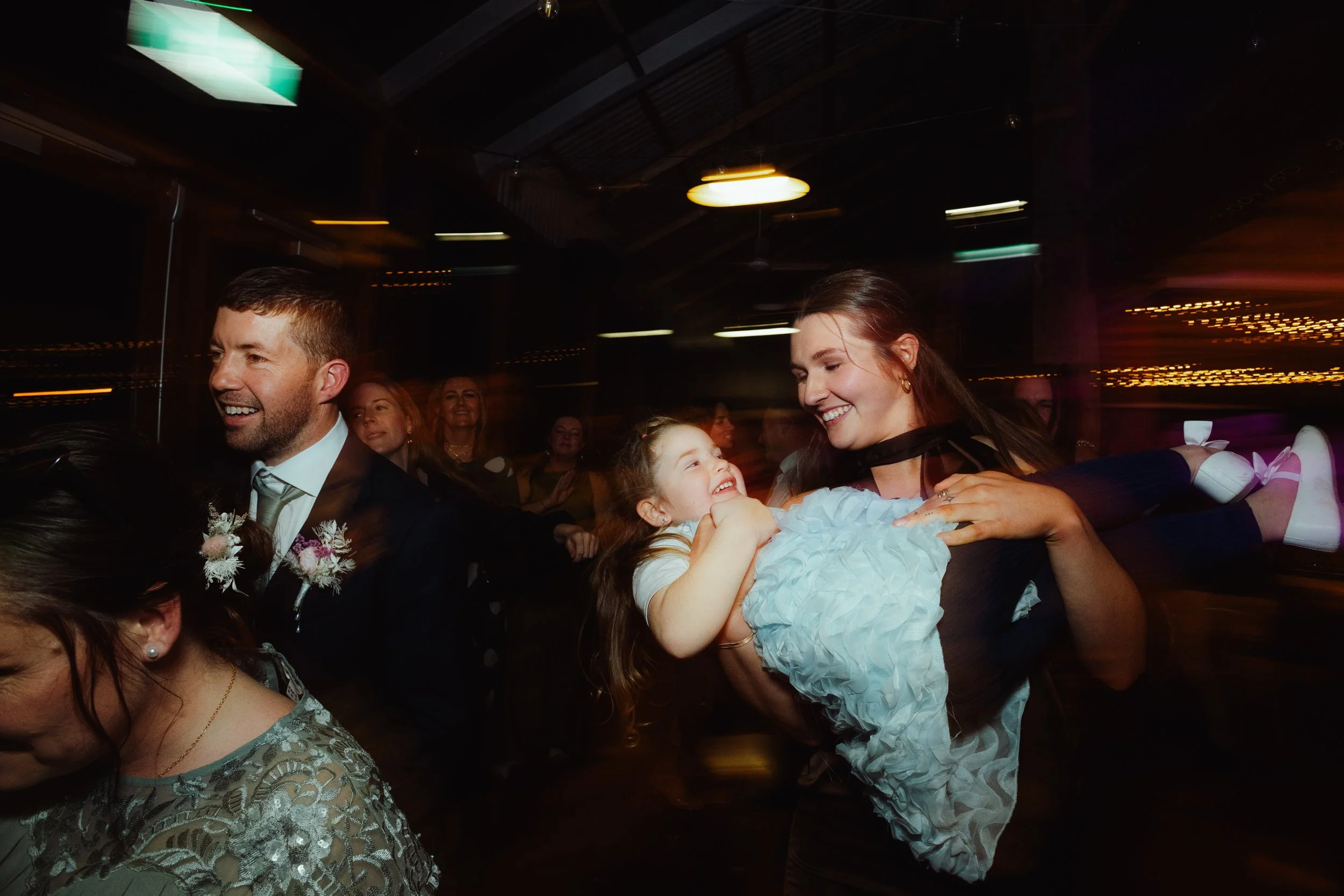 A joyful scene of a group of people at a celebration or party inside a dimly lit venue. A woman holding a young girl smiles at each other, surrounded by other guests. The atmosphere is lively with blurred lights indicating movement and excitement.