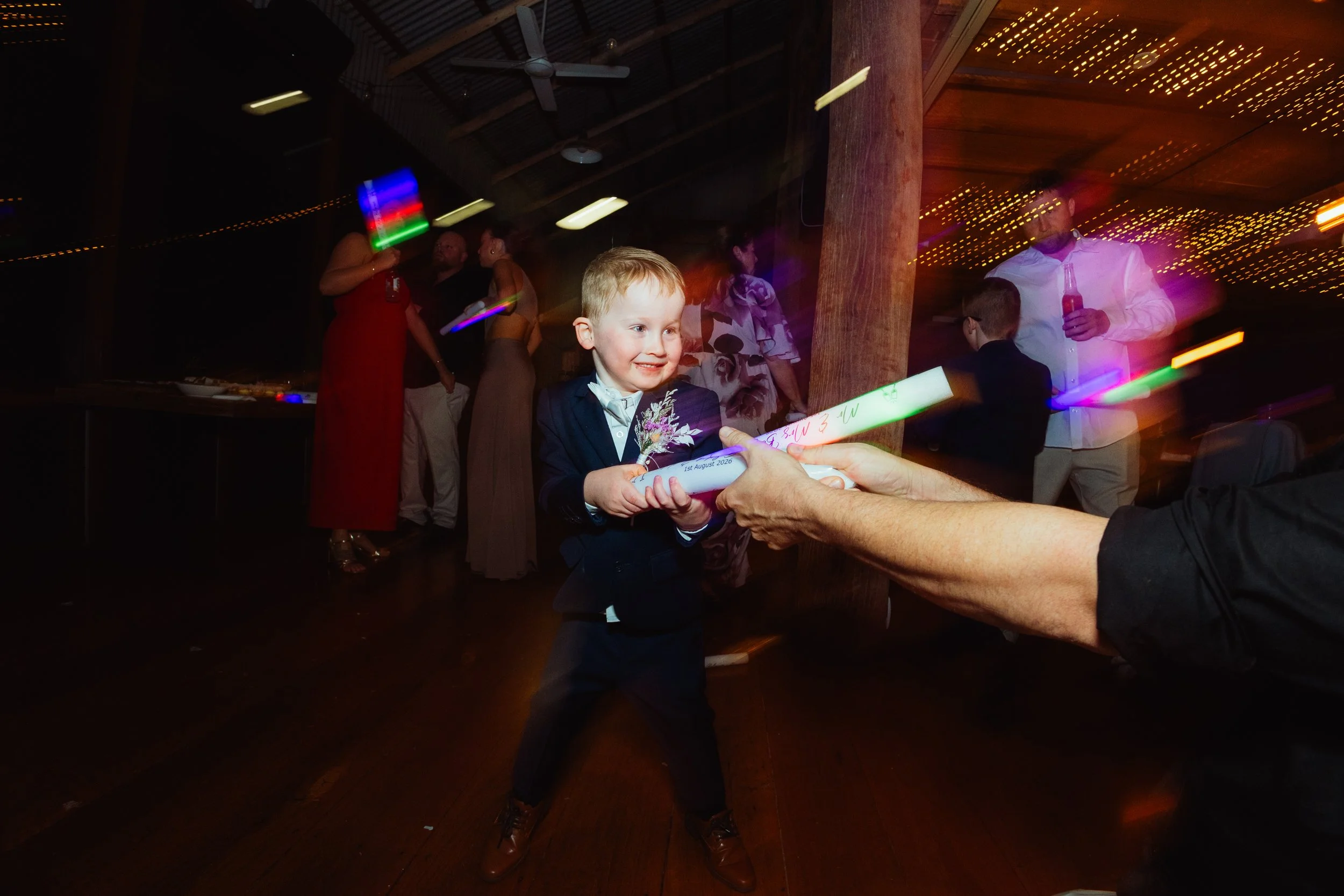 A young boy in a tuxedo receiving a rolled scroll with a birthday message at a celebration with adults in the background, colorful lights, and a rustic wooden interior.