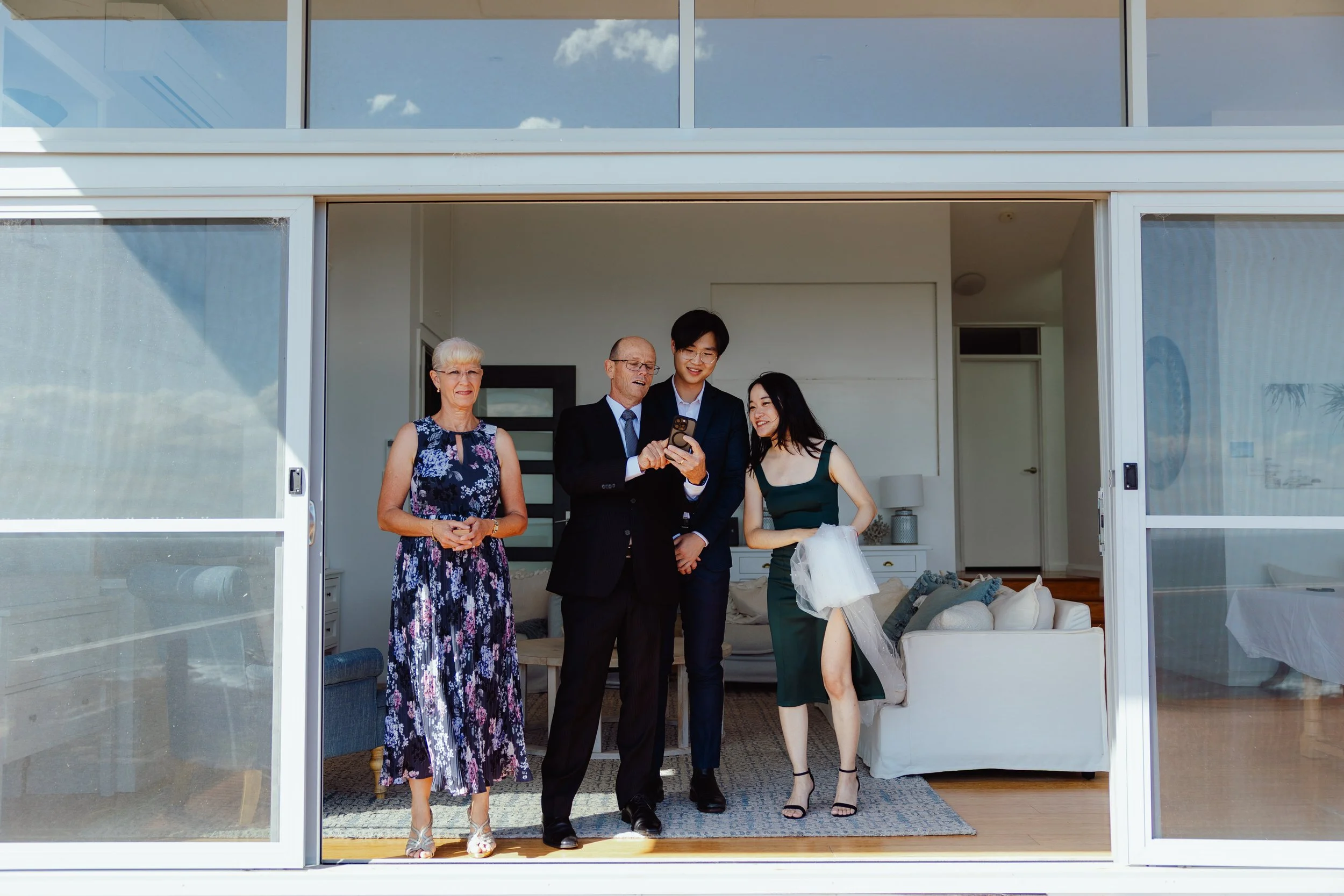 Four people standing inside a room with open sliding glass doors, looking at a phone and smiling. Two women and two men, dressed in semi-formal attire, with a living room visible behind them.