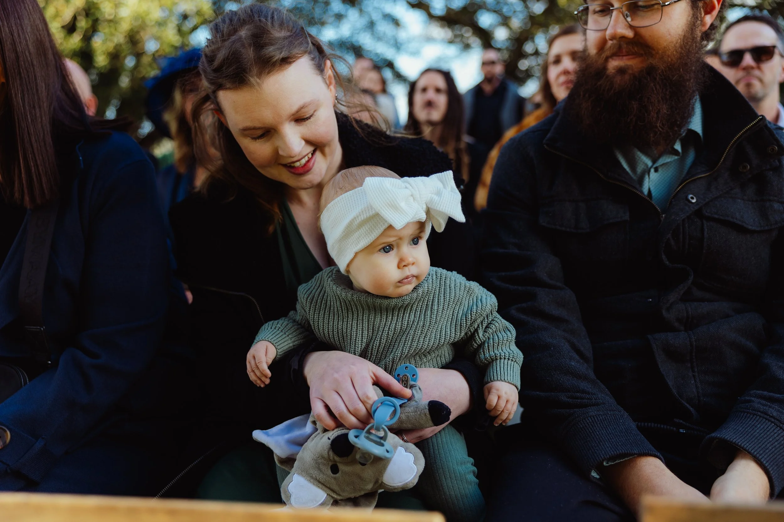 A young woman with light brown hair holding a baby with blue eyes and a beige sun hat, sitting outdoors among a crowd of people, some wearing sunglasses, in a park with green trees in the background.