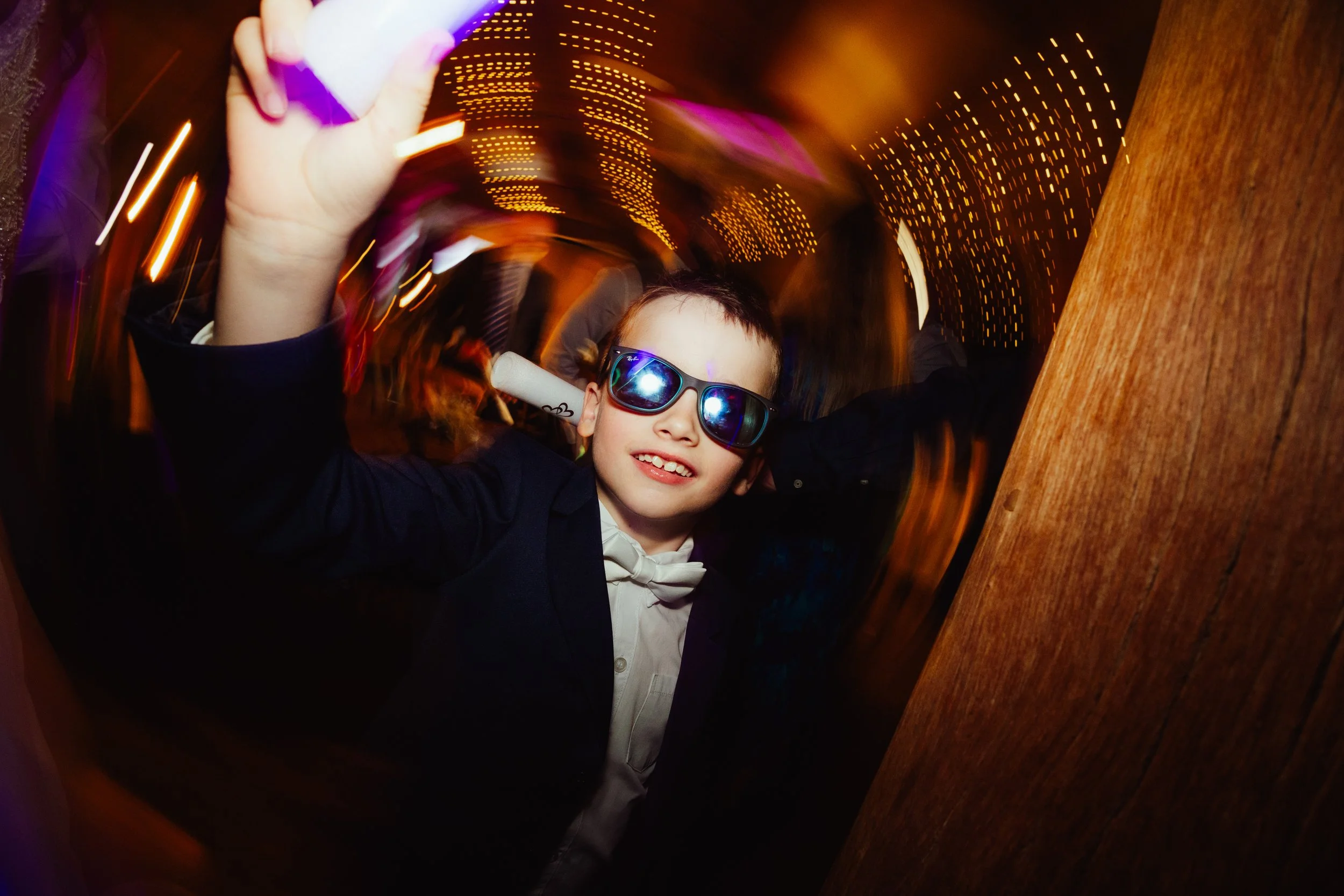 A young boy wearing sunglasses and a tuxedo with a bowtie, smiling at the camera, with blurred colorful lights in the background, suggesting a lively and energetic environment.