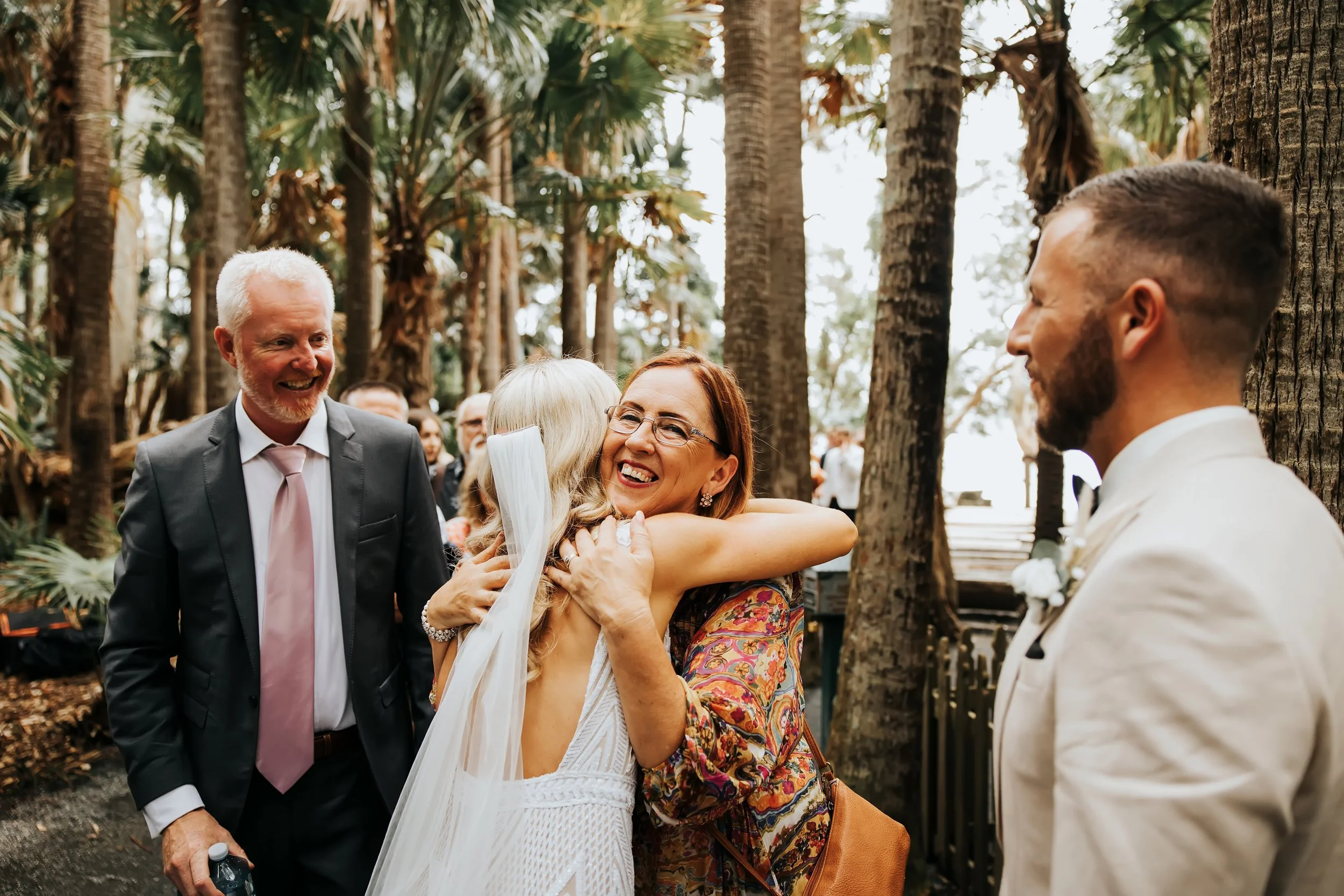 A group of people at a wedding ceremony outdoors among tall trees. A woman in a white dress is hugging an older woman, while a man with a beard and a suit looks on smiling.