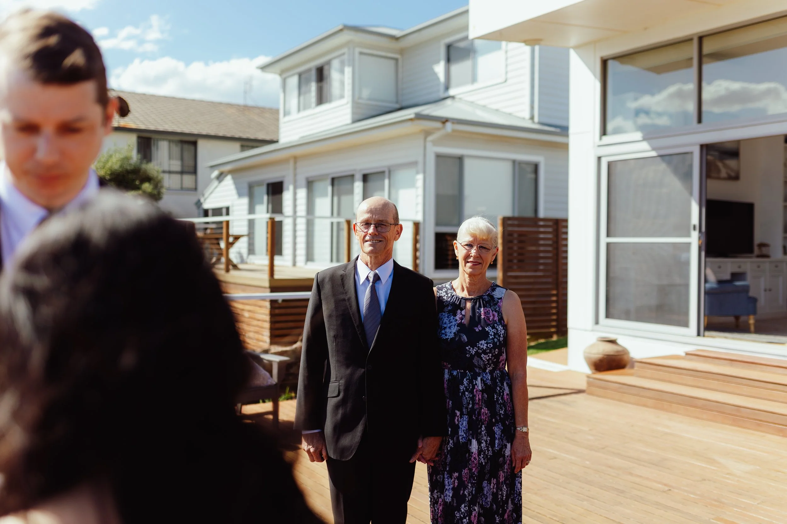 A group of people standing outdoors in front of modern white houses with large windows. The focus is on an elderly couple, a man in a suit and a woman in a floral dress, smiling at the camera. A blurred person is in the foreground on the left. The sk