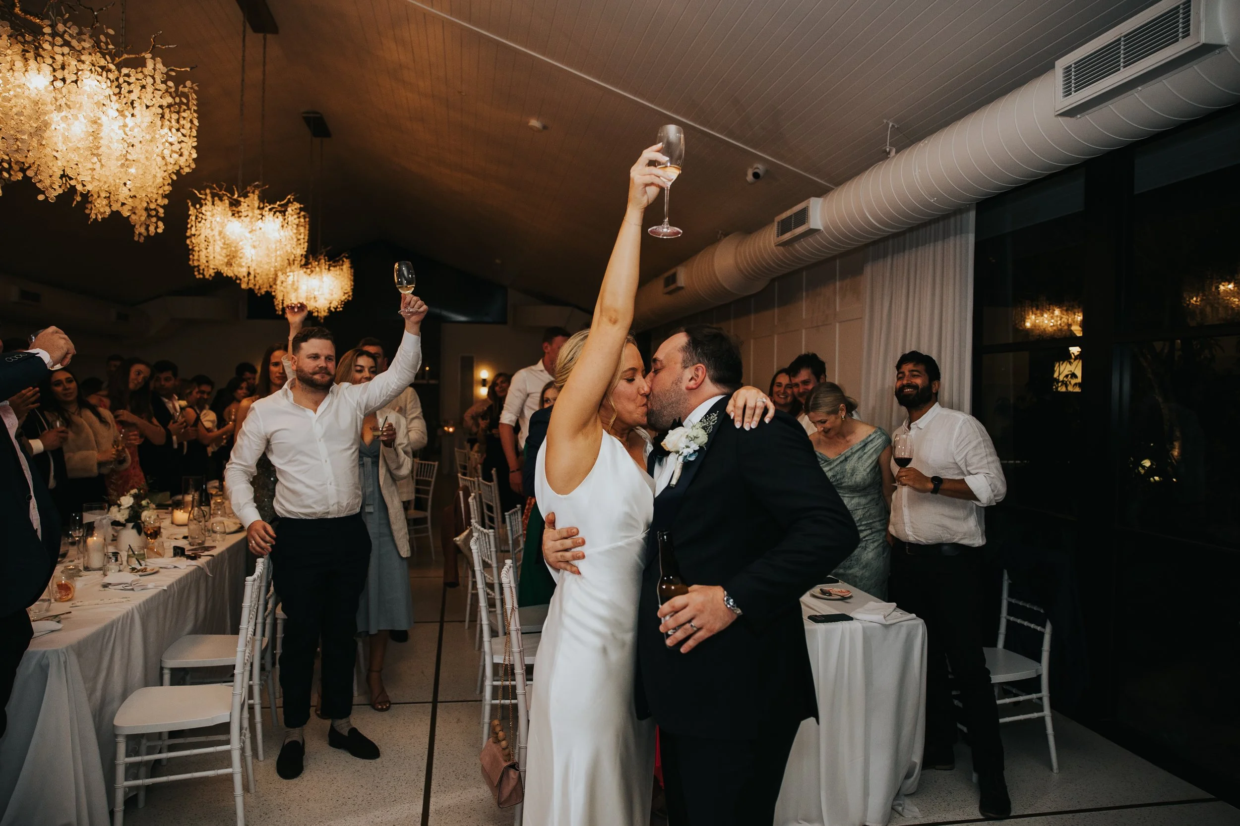 A newly married couple sharing a kiss at their wedding reception, surrounded by guests celebrating and raising glasses in a decorated indoor venue with chandeliers.