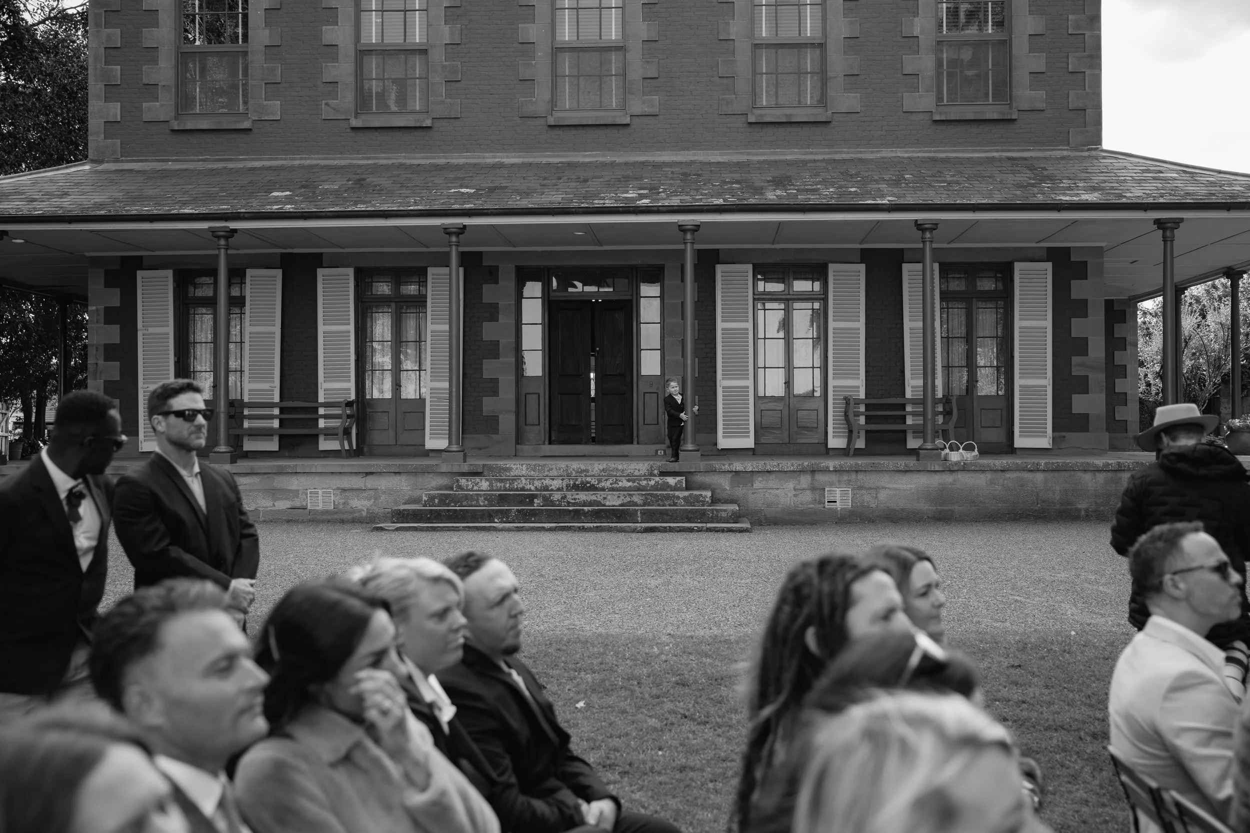 A black and white photo of a large brick house with a porch and multiple windows. Several people are seen in the foreground, seated or standing, with a woman standing near the porch door.