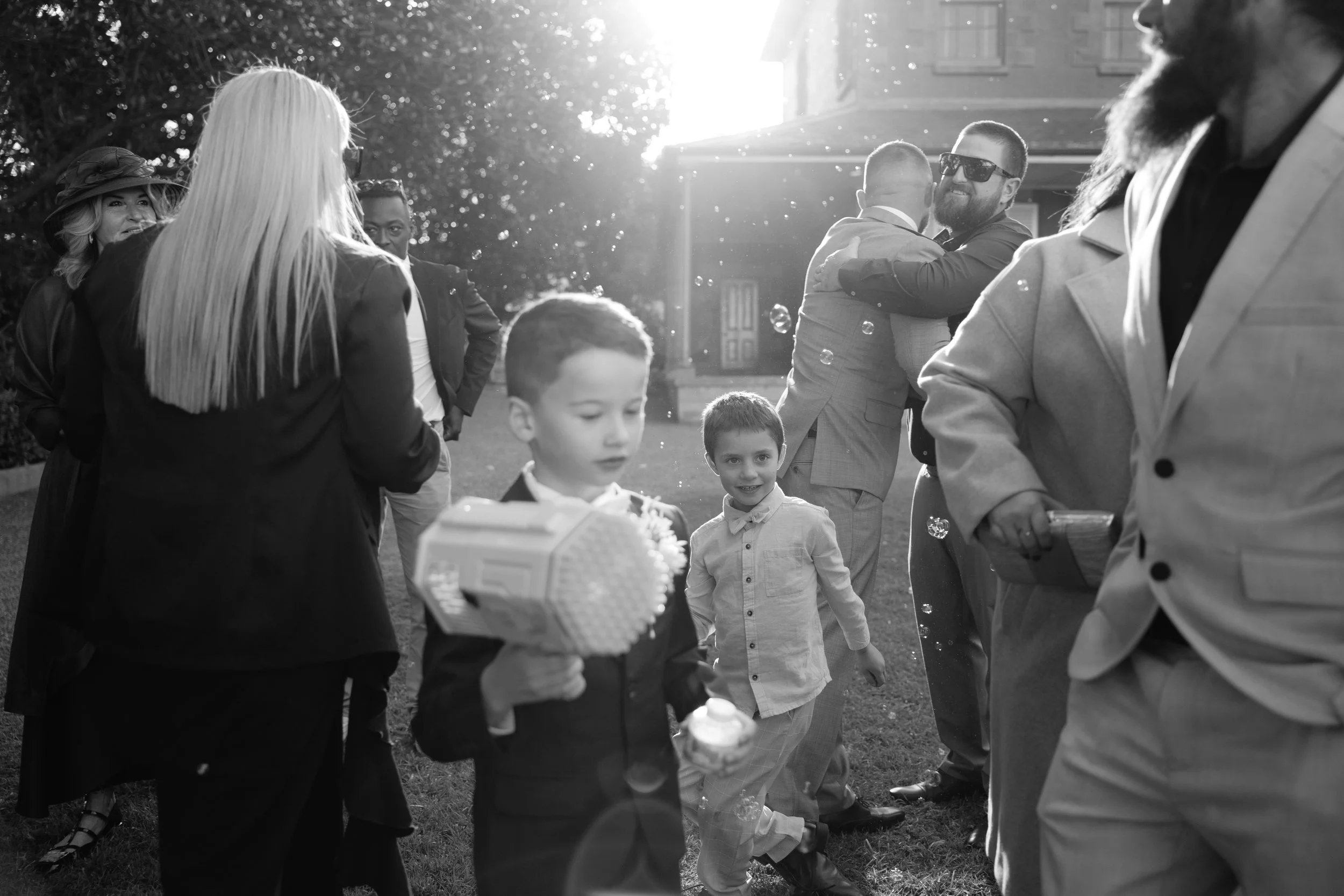 People at an outdoor gathering, including children and adults, with some embracing, around bubbles, on a sunny day with a house in the background.